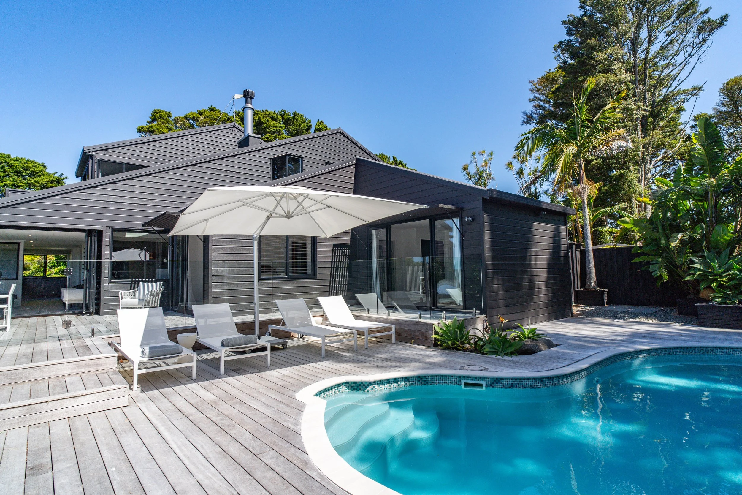 Modern house with a dark exterior, outdoor pool, lounge chairs, and white umbrella on a wooden deck, surrounded by tall trees and clear blue sky.