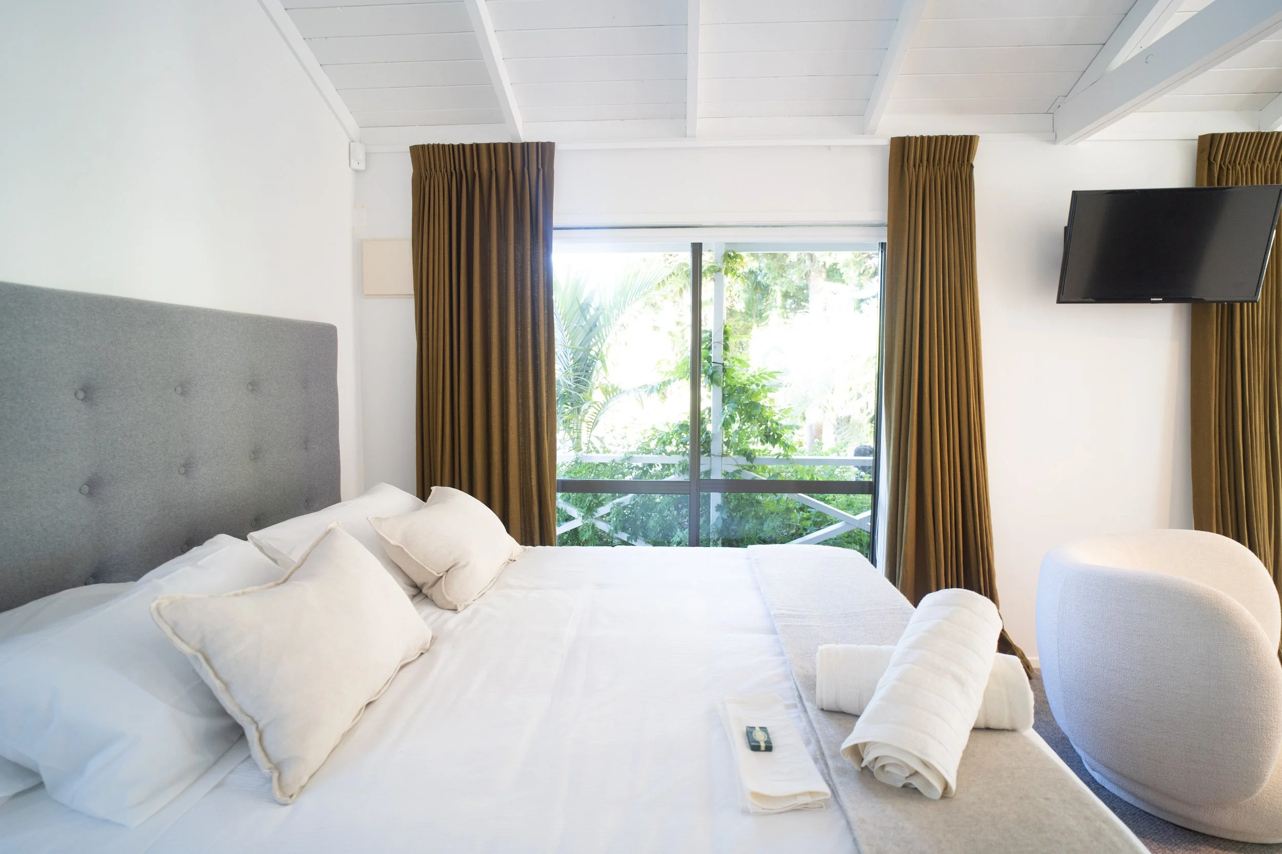 A bedroom with a large bed covered in white linens and multiple pillows, a gray upholstered headboard, a sliding glass door with brown curtains, and a wall-mounted flat-screen TV, with greenery visible outside.