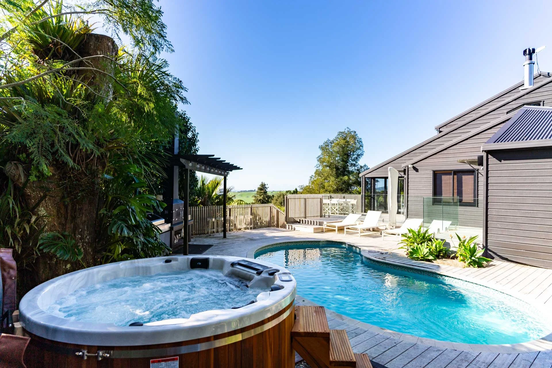 Backyard with a swimming pool, hot tub, lounge chairs, deck, wooden fencing, and house with gray siding and solar panels on the roof. Trees and blue sky in the background.