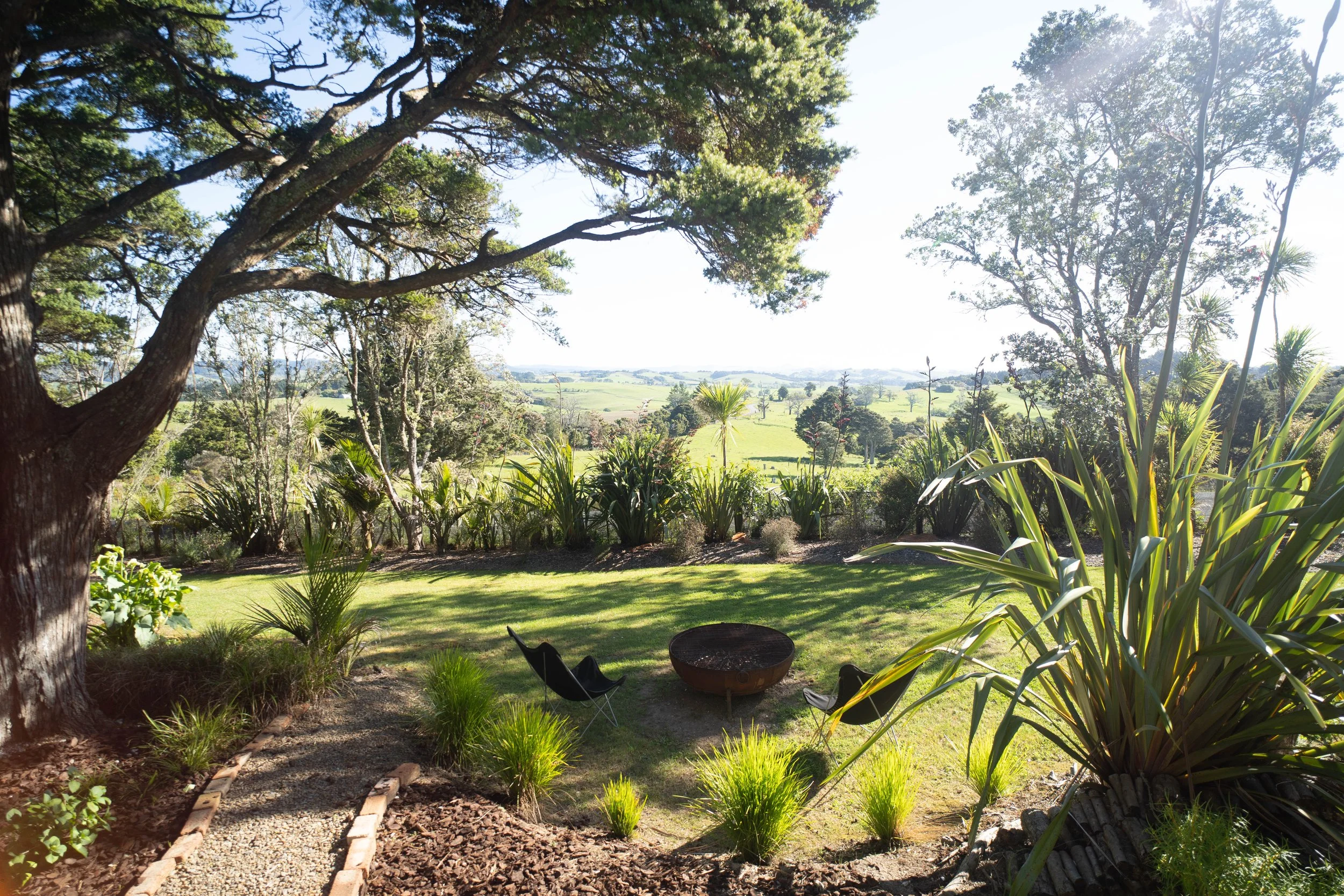 A spacious backyard garden with lush green grass, various plants, and tall trees under a bright sky. There are black lounge chairs and a firepit in the yard, with a scenic view of rolling hills in the background.