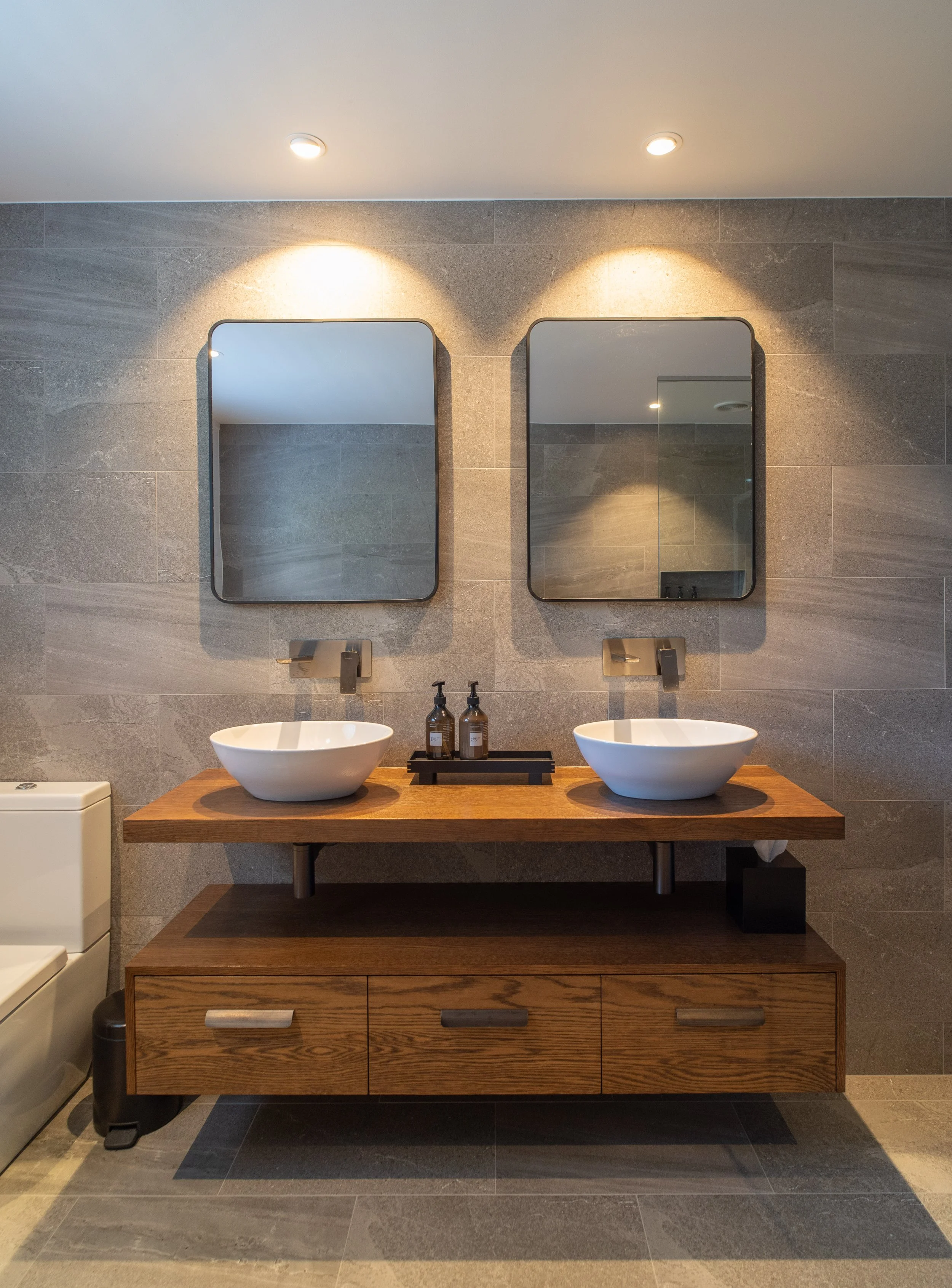 Modern bathroom with two rectangular mirrors, two vessel sinks, a wooden vanity cabinet, and two soap dispensers on a wooden tray. The walls are tiled in gray, and the ceiling has two recessed lights.