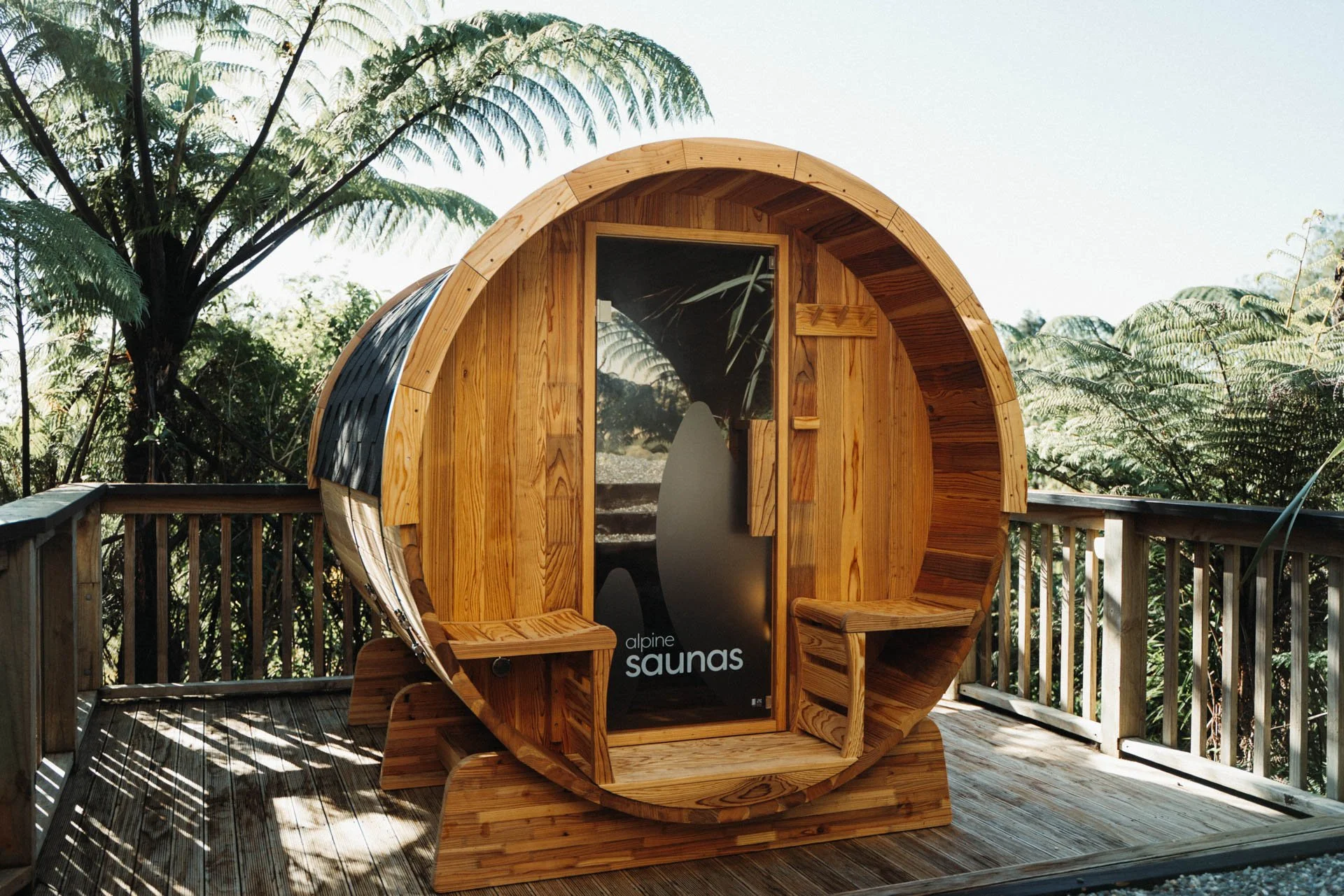 A wooden barrel-shaped sauna on a outdoor deck surrounded by trees and greenery.