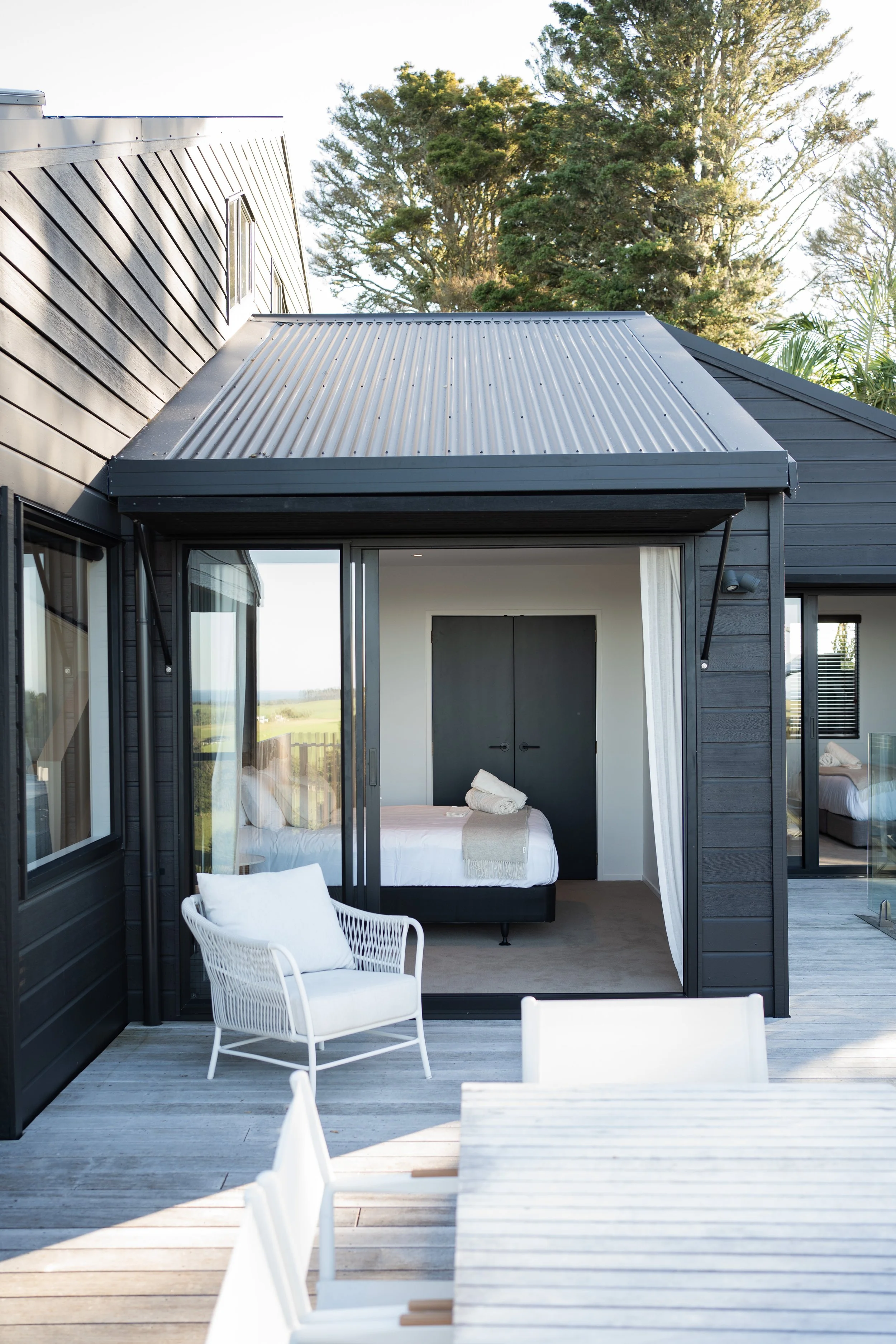 Modern house with black exterior walls and metal roof, featuring an open bedroom with white bedding, sitting area with white chairs on a wooden deck, large trees in the background.