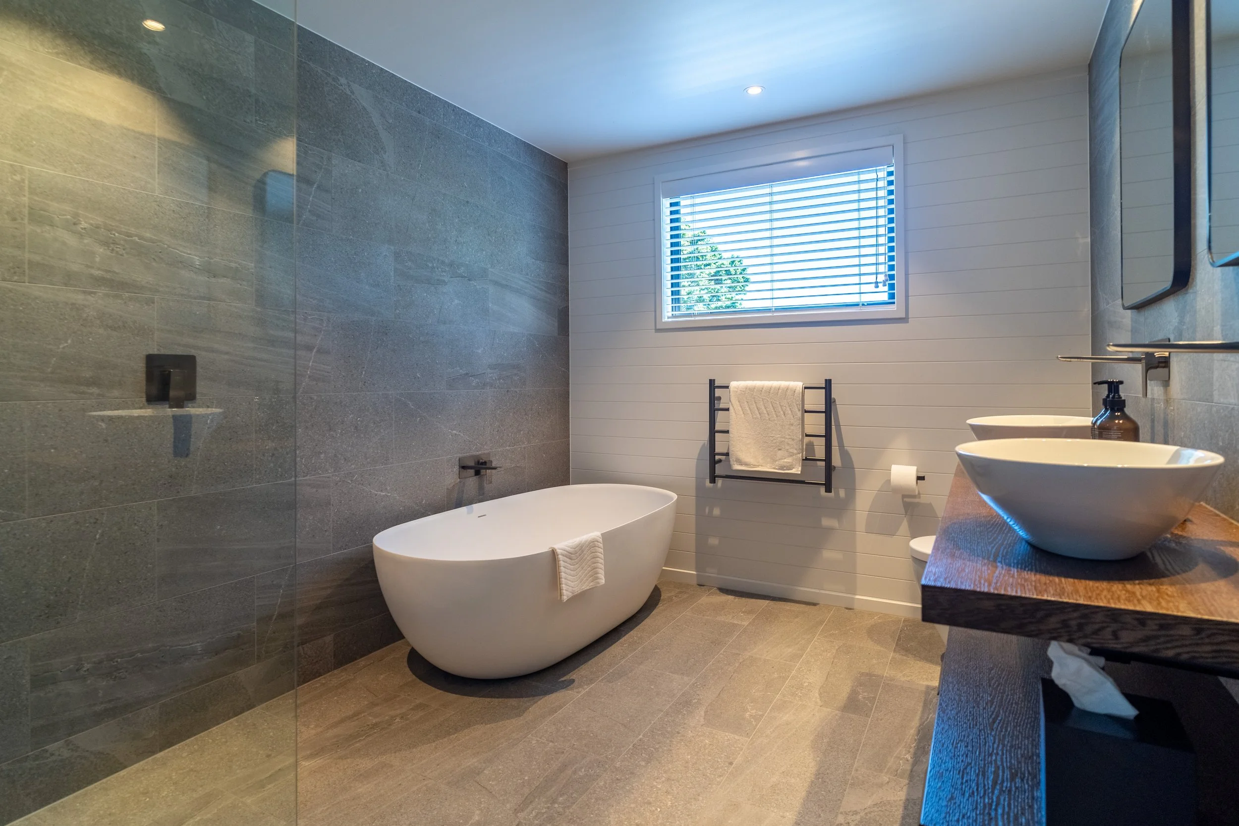 Modern bathroom with gray tiled shower area, freestanding bathtub, white wood-paneled wall, rectangular window with blinds, double sinks with brown countertop, brown soap dispenser, and towel rack with a white towel.
