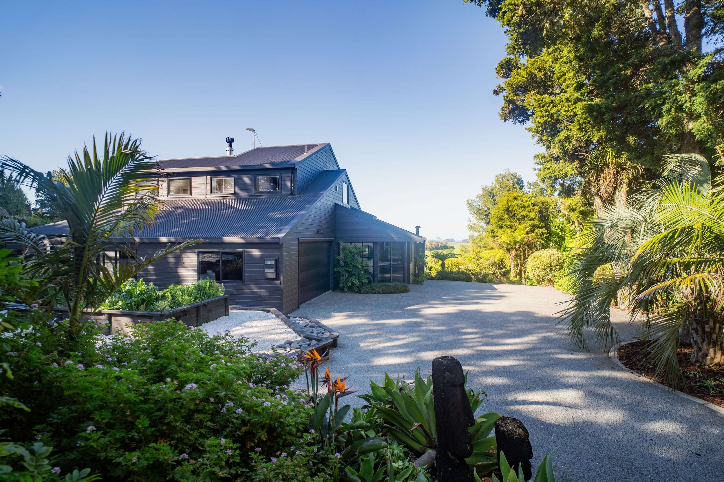 A modern house with dark exterior siding, surrounded by lush greenery and trees, with a gravel pathway and landscaped garden.