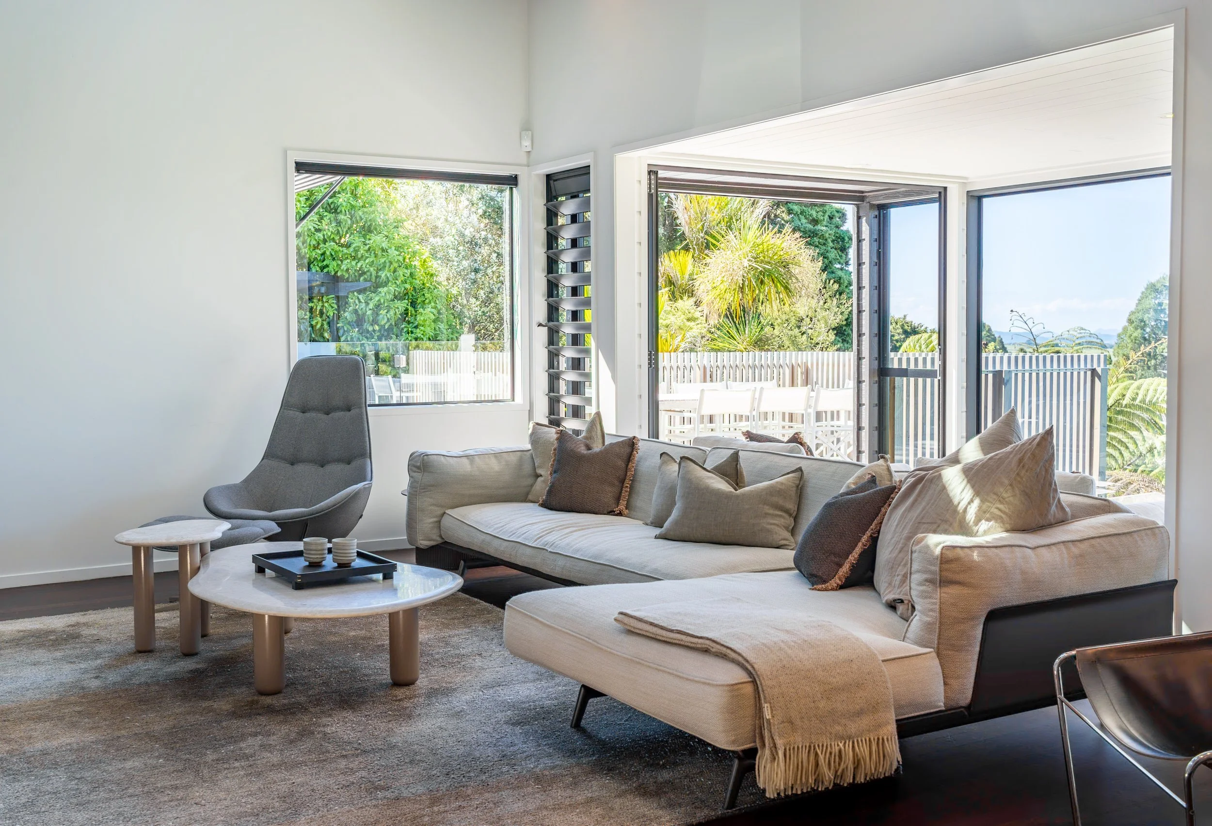 Living room with beige sofa and pillows, gray armchair, two round coffee tables, large windows showing outdoor greenery and sky.