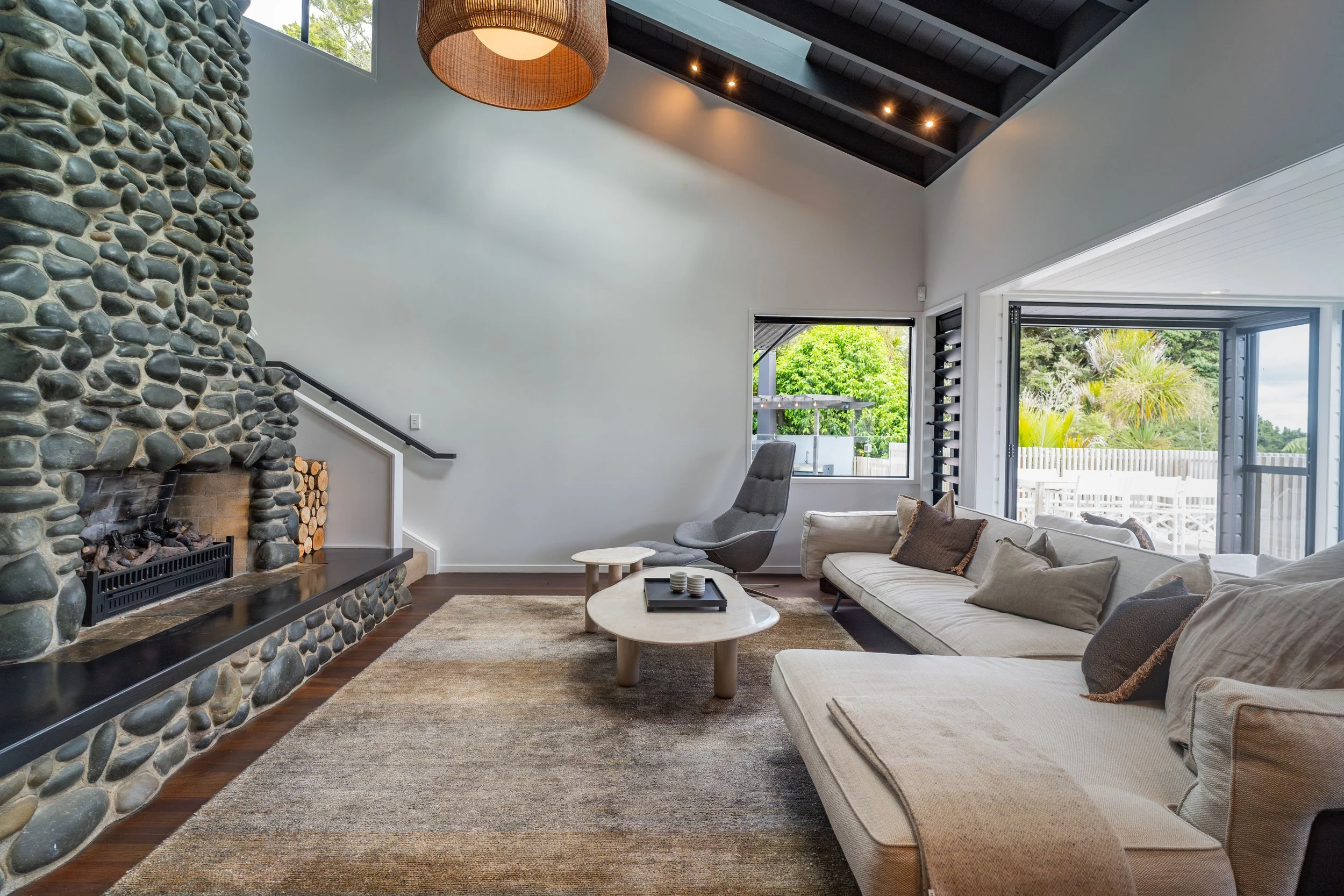 Modern living room with a large stone fireplace, a beige sectional sofa, a gray armchair, a small round coffee table, and large windows revealing greenery outside.
