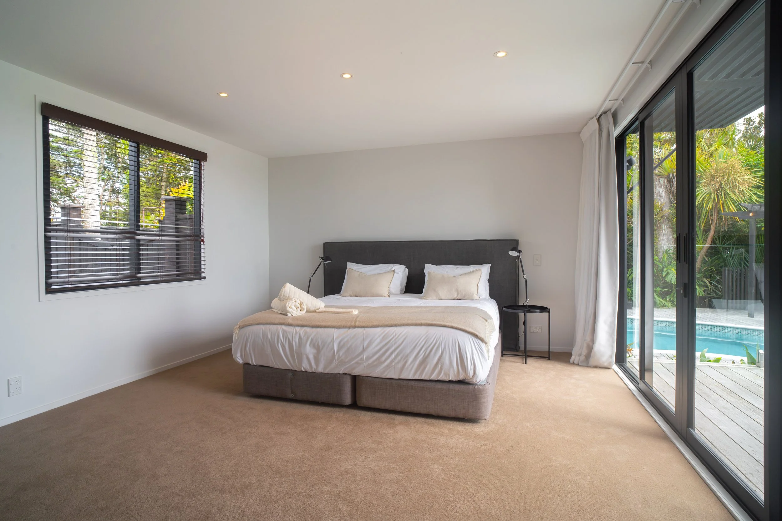 Bedroom with beige carpet, white walls, large window with wooden blinds, bed with gray headboard, white bedding, and beige pillows, sliding glass door to patio with greenery outside.