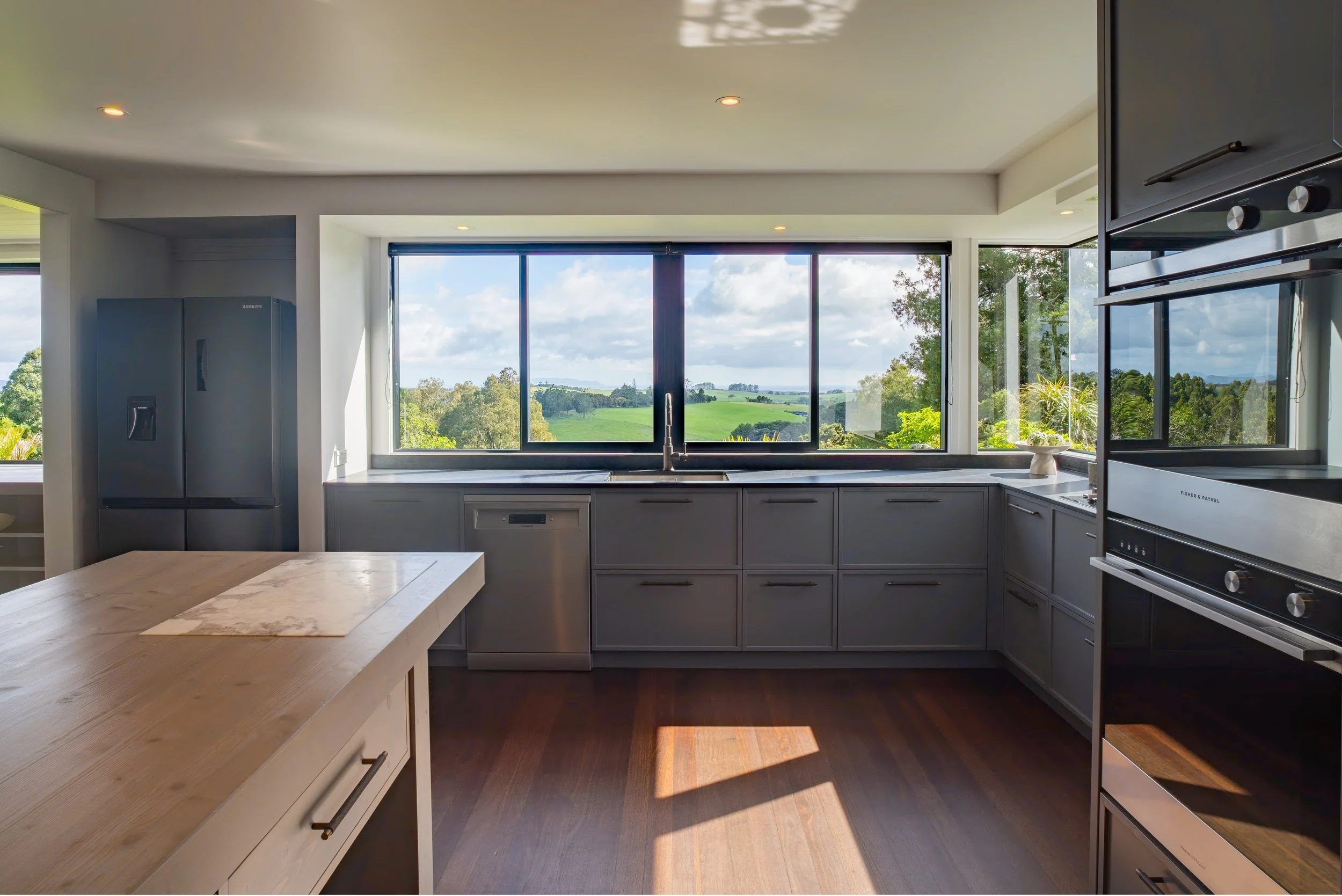 Modern kitchen with large window overlooking green fields and trees, gray cabinetry, stainless steel appliances, and a wooden dining table.