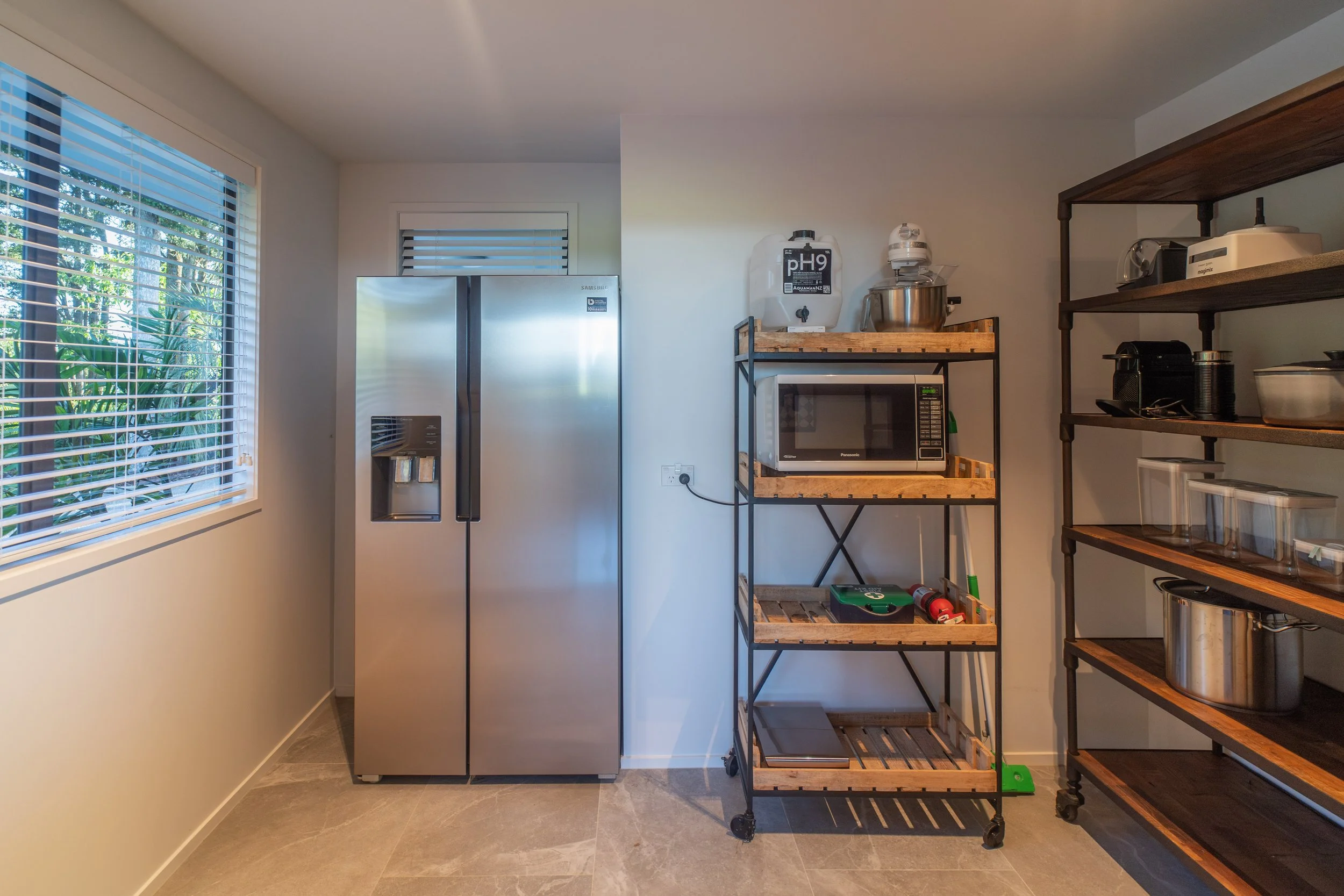 A kitchen corner with a large stainless steel refrigerator on the left, and on the right, a black metal and wood shelving unit with a microwave oven, cleaning supplies, and various kitchen appliances. A wooden open shelf to the right contains more ki