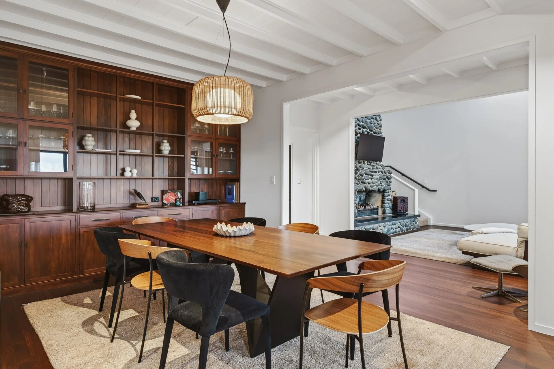 A dining room with a large wooden table surrounded by black and wooden chairs, a wooden built-in cabinet with decorative white vases, a hanging wicker light fixture, and a living room with a stone fireplace and a television in the background.