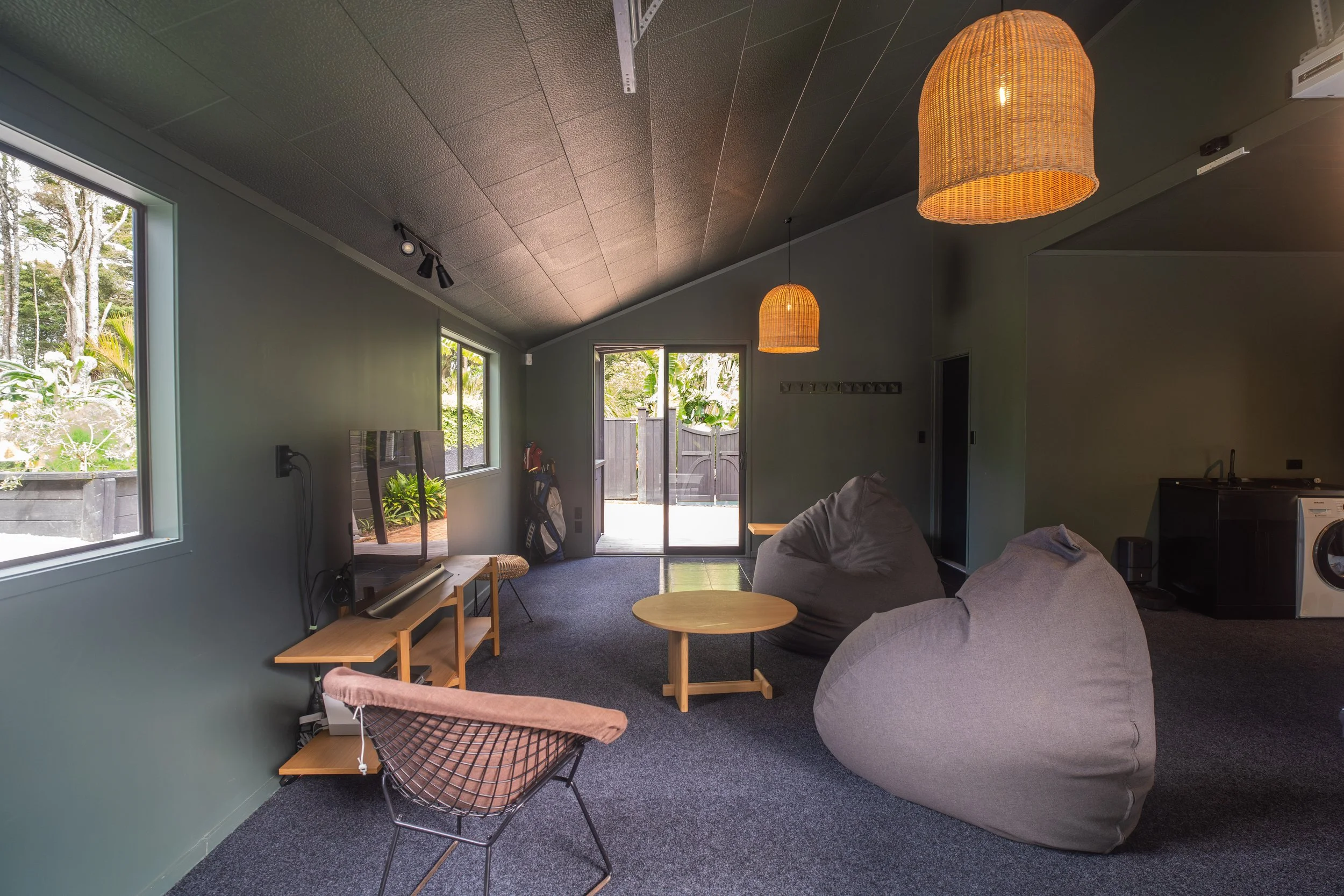 Living room with large window and glass door, gray walls, dark ceiling, hanging wicker lamps, gray sofa, wooden side tables, and a TV, with outdoor greenery visible.