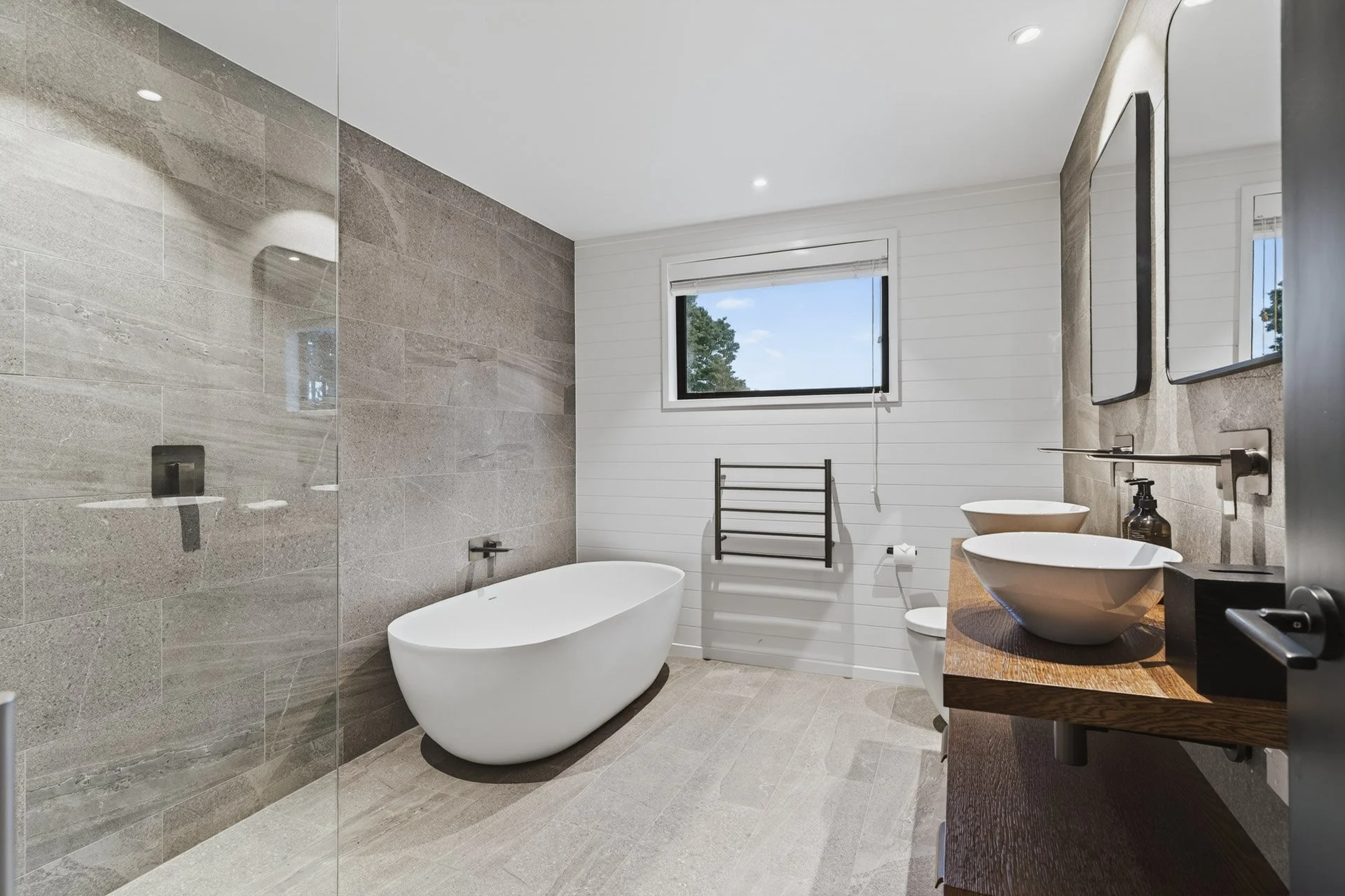 Modern bathroom with gray tiled walls and floor, white bathtub, two vessel sinks on wooden vanity, black faucet and accessories, black towel rack, window showing trees and blue sky, ceiling lights.