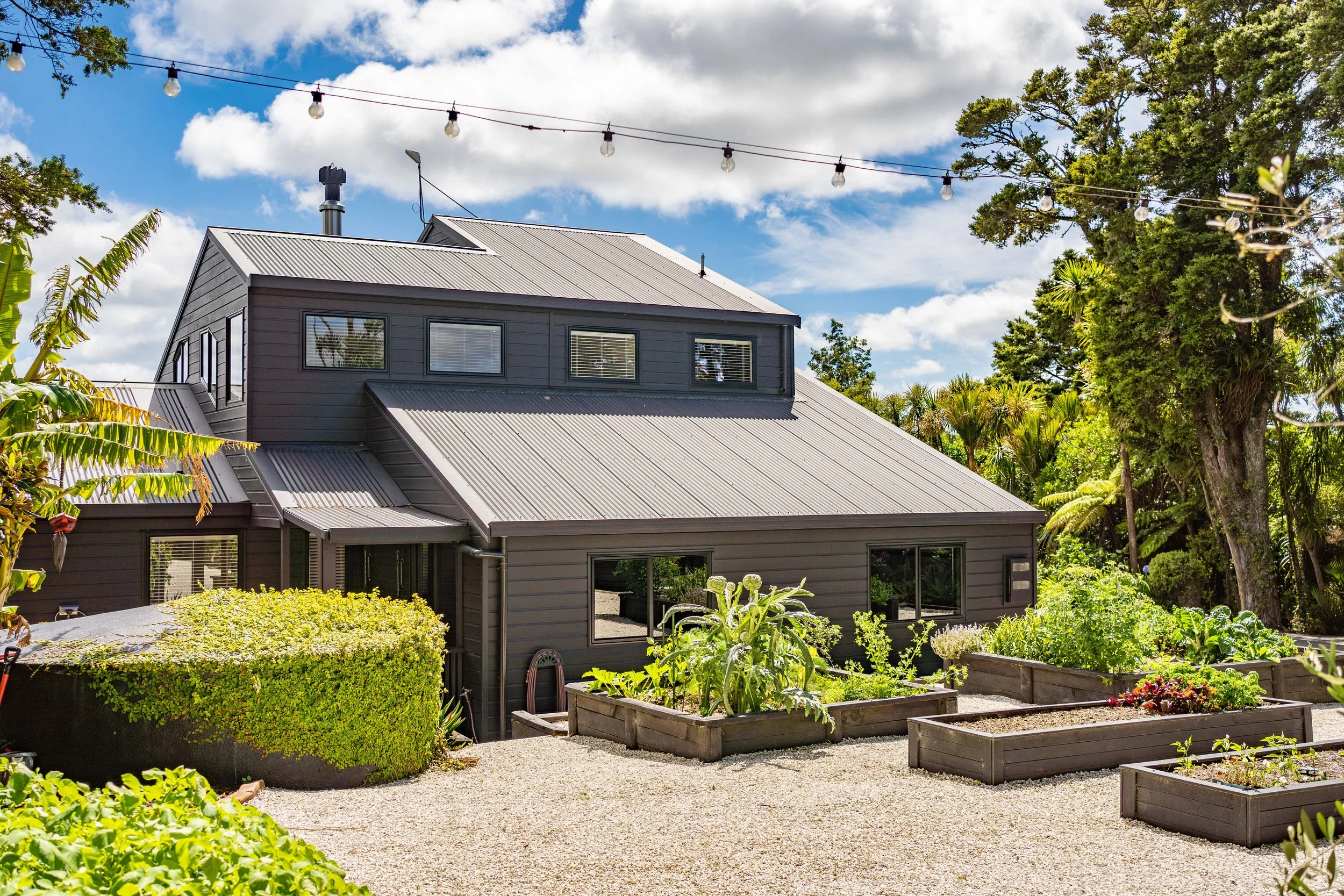 A gray house with a metal roof surrounded by a lush vegetable garden and trees, under a blue sky with scattered clouds.