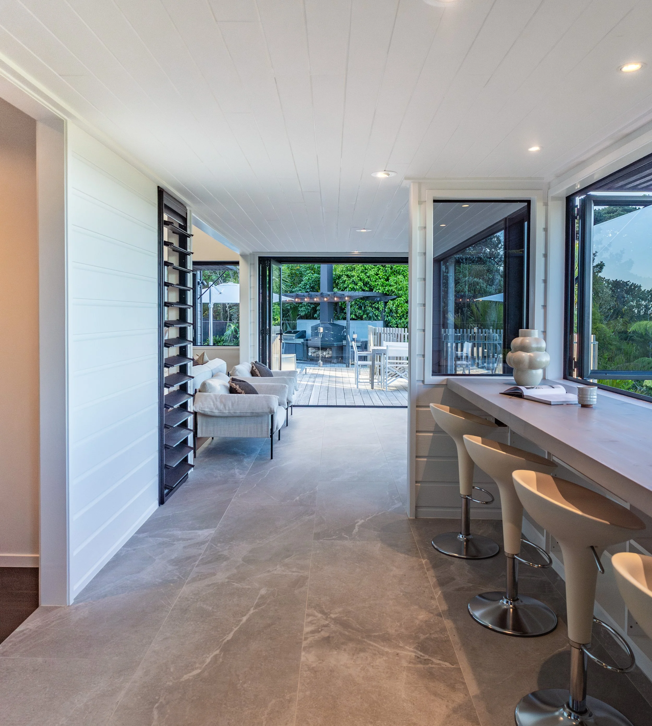 Modern indoor living space with open glass doors leading to an outdoor patio area with patio furniture and greenery. The room features a gray tiled floor, white walls, a white ceiling with recessed lighting, and a kitchen bar area with white bar stoo