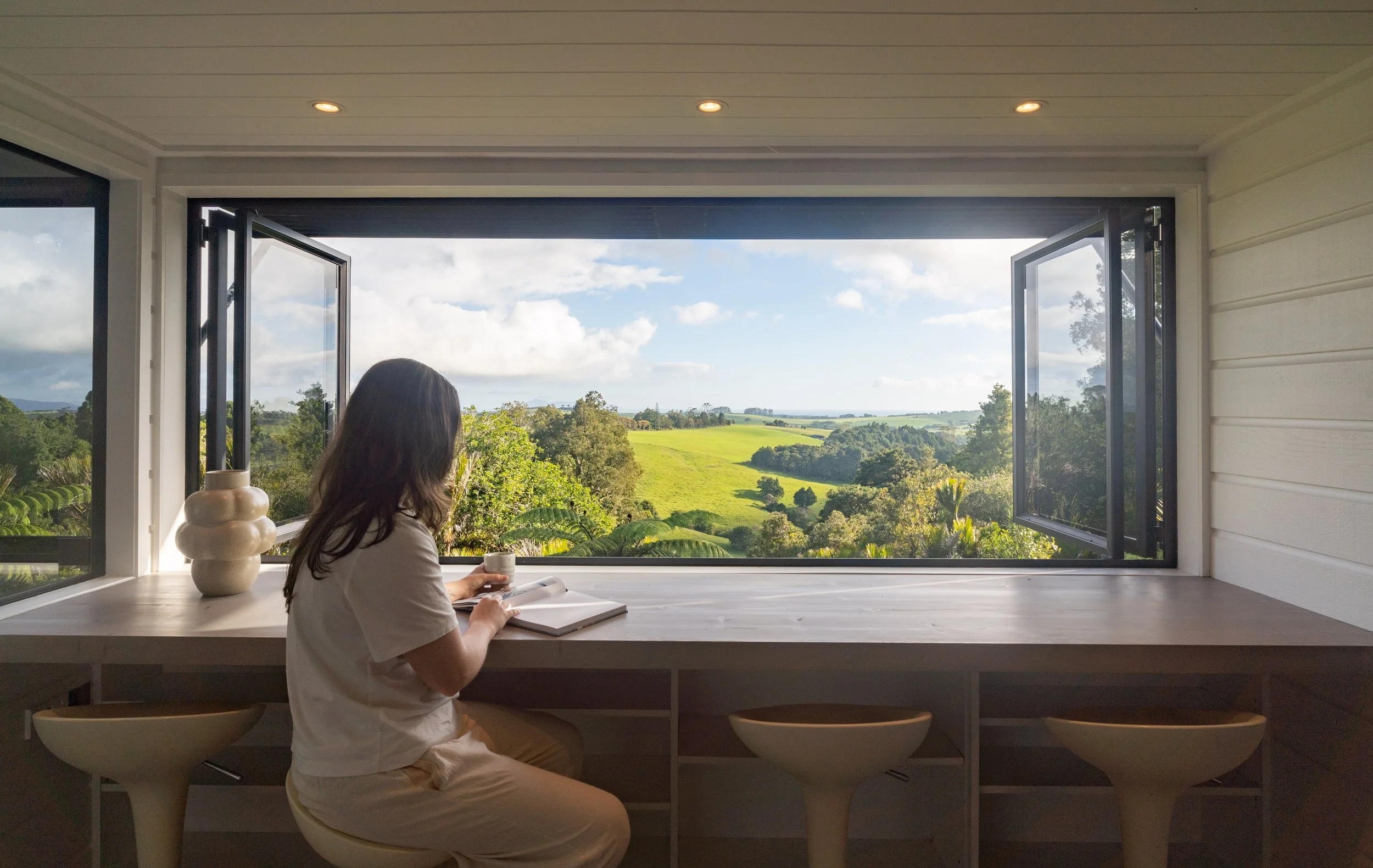 A woman sitting at a white counter, reading a book and holding a cup, with an open window showing green fields and trees outside.