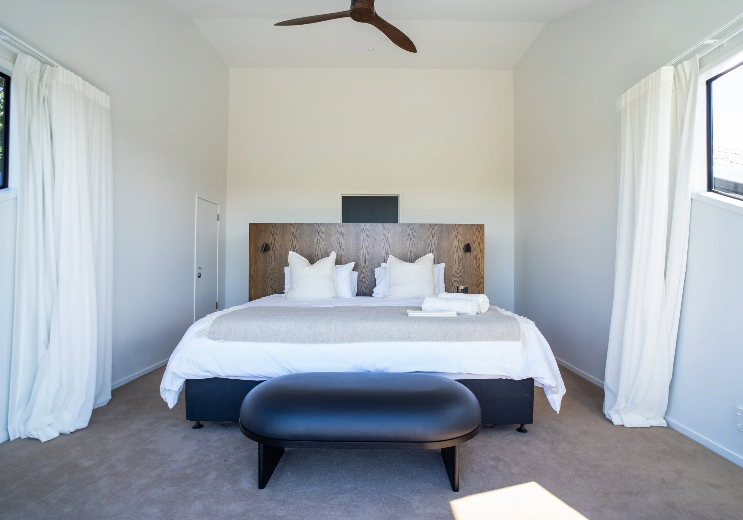 A bedroom with a neatly made bed featuring white pillows and linens, a wooden headboard, black reading lamps on either side, and large windows with white curtains, a ceiling fan above, and a small black bench at the foot of the bed.