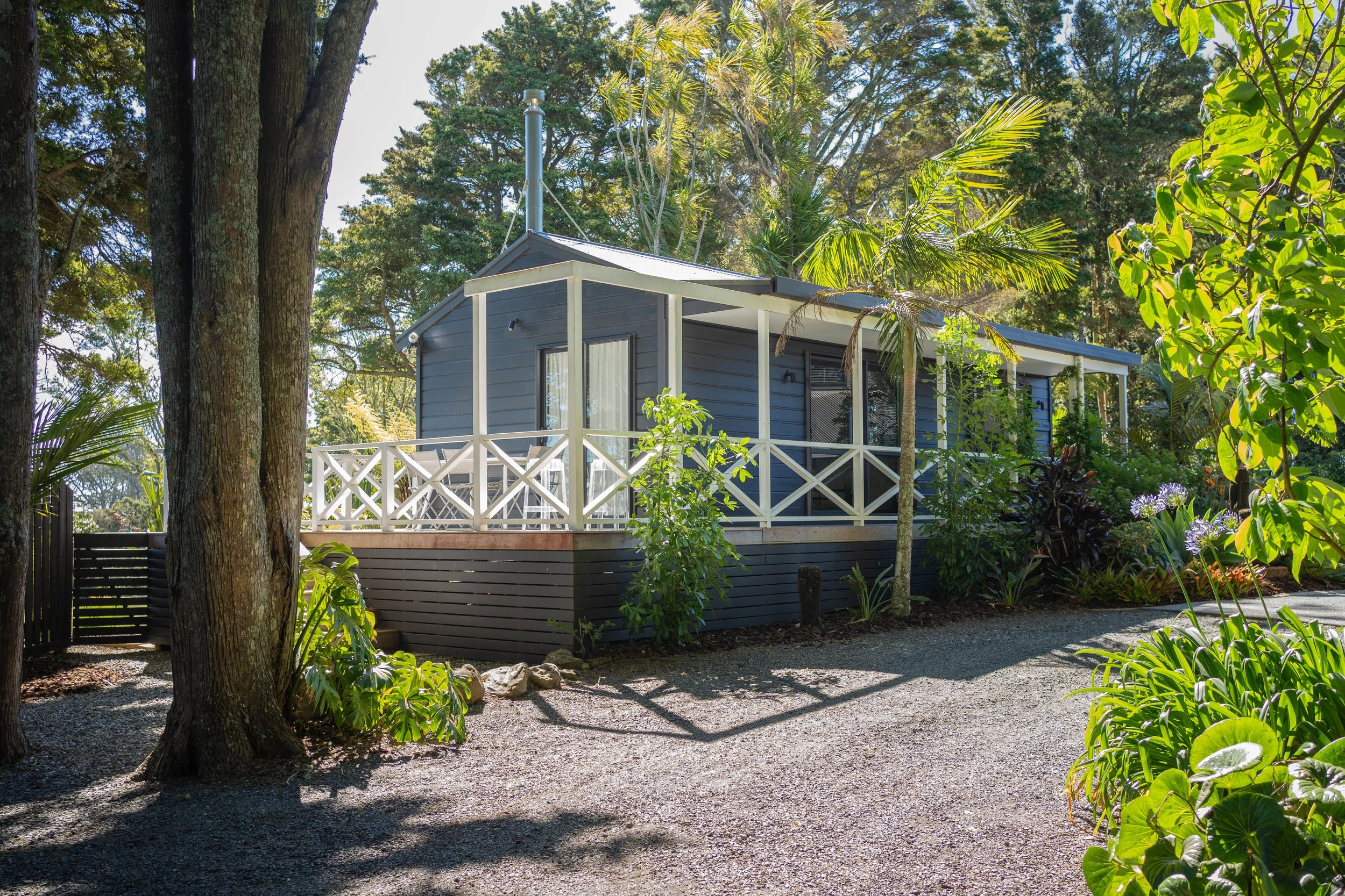 A small house or cottage painted in dark gray with white trim, situated in a lush garden surrounded by tall trees, green plants, and colorful flowers. There is a deck with white railing, and a gravel pathway leading to the house, with sunlight filter