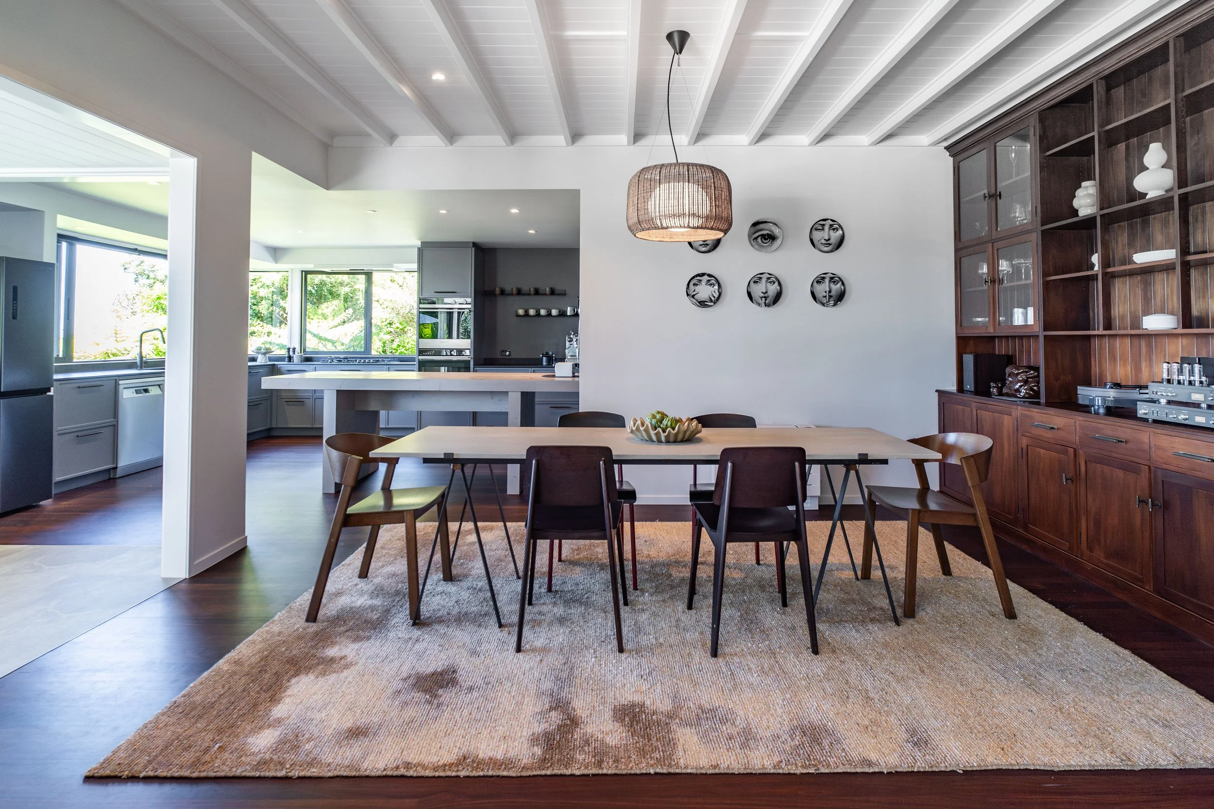 A modern dining area with a wooden table and black chairs, an area rug, and a large wooden cabinet with white decorative items, in an open kitchen space with large windows showing greenery outside.