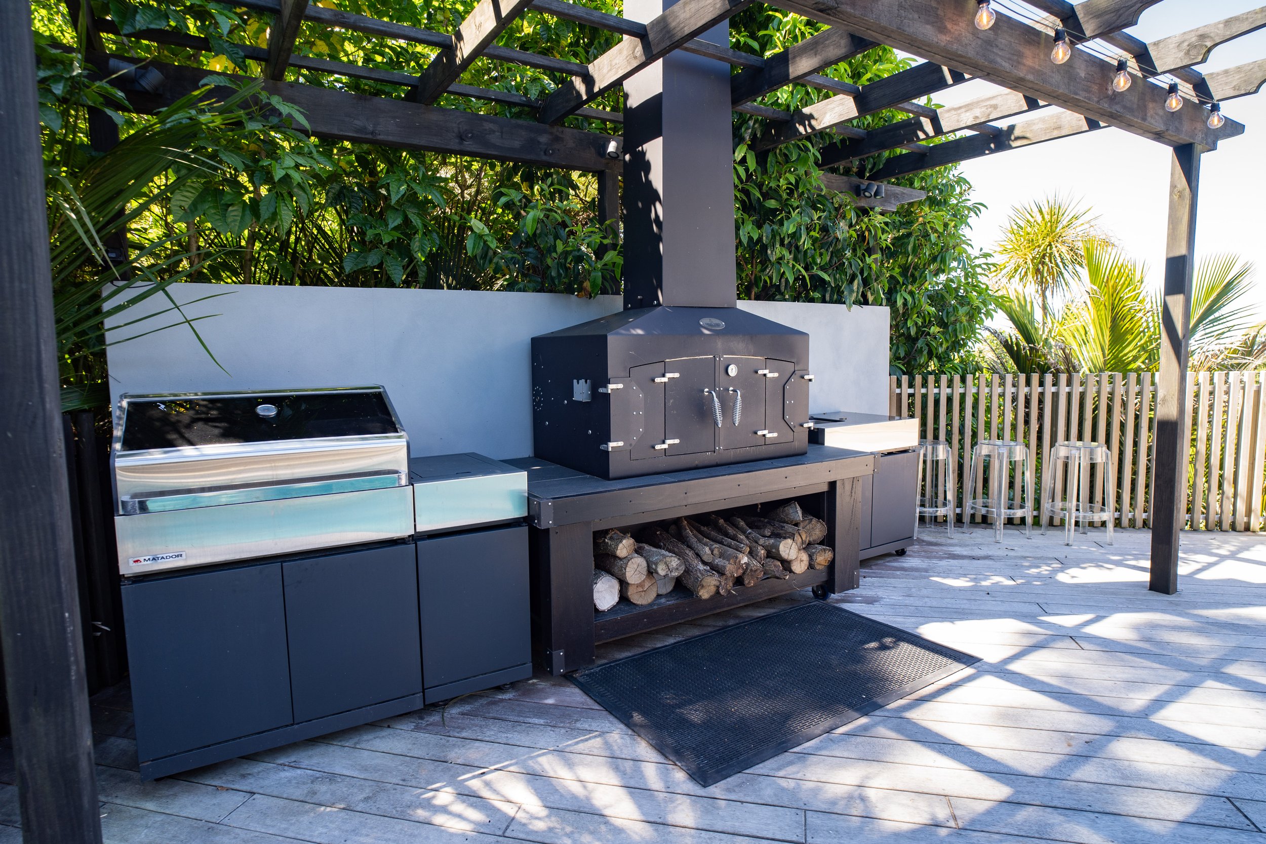 Outdoor BBQ area with a black grill, wood storage underneath, a stainless steel grill on the left, and four acrylic barstools on a wooden deck, surrounded by lush green foliage and a fence, under string lights.
