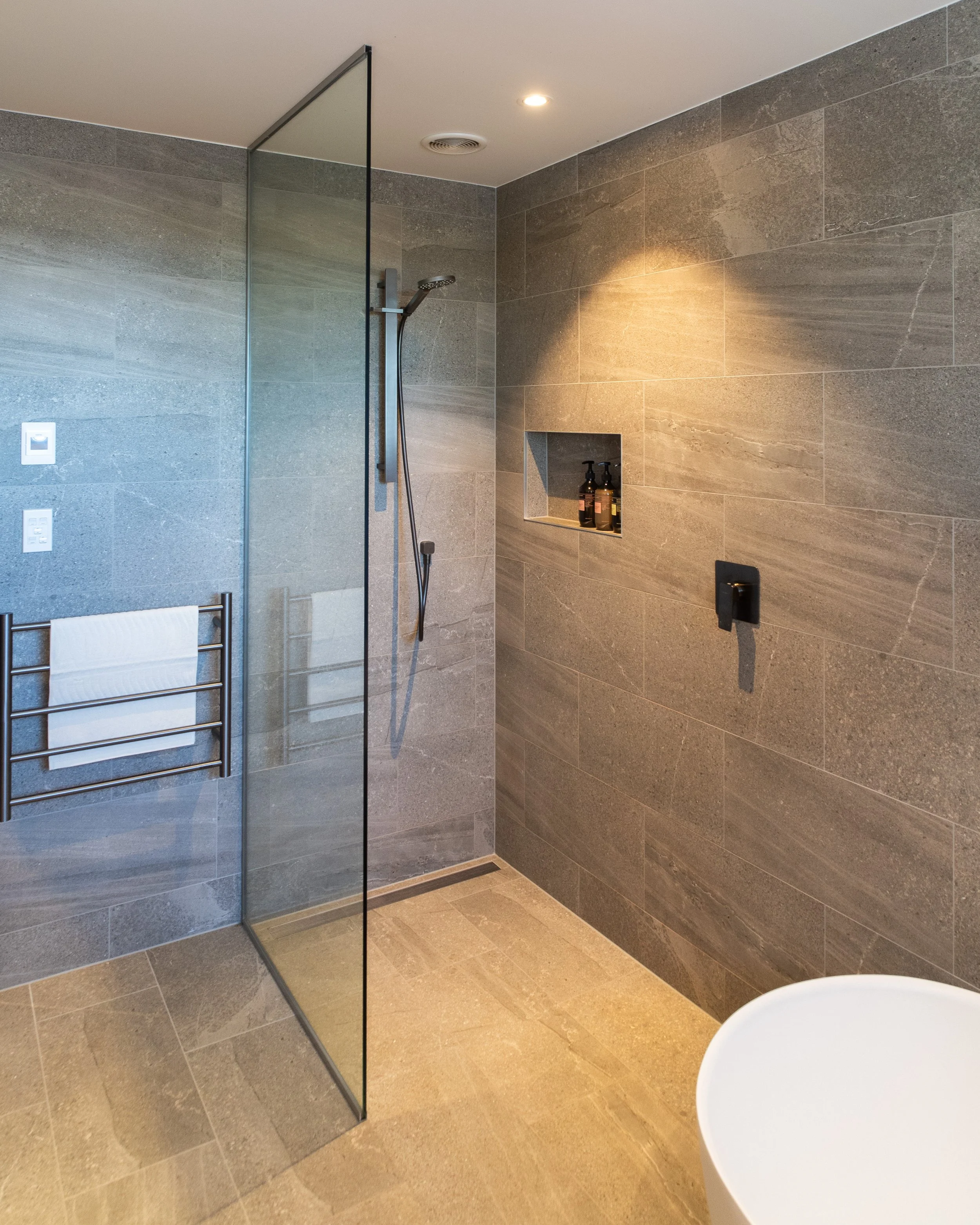 Modern bathroom shower with gray stone tiles, a wall-mounted showerhead, a built-in shelf with shampoo bottles, a glass partition, a heated towel rack, and a white bathtub in the corner.