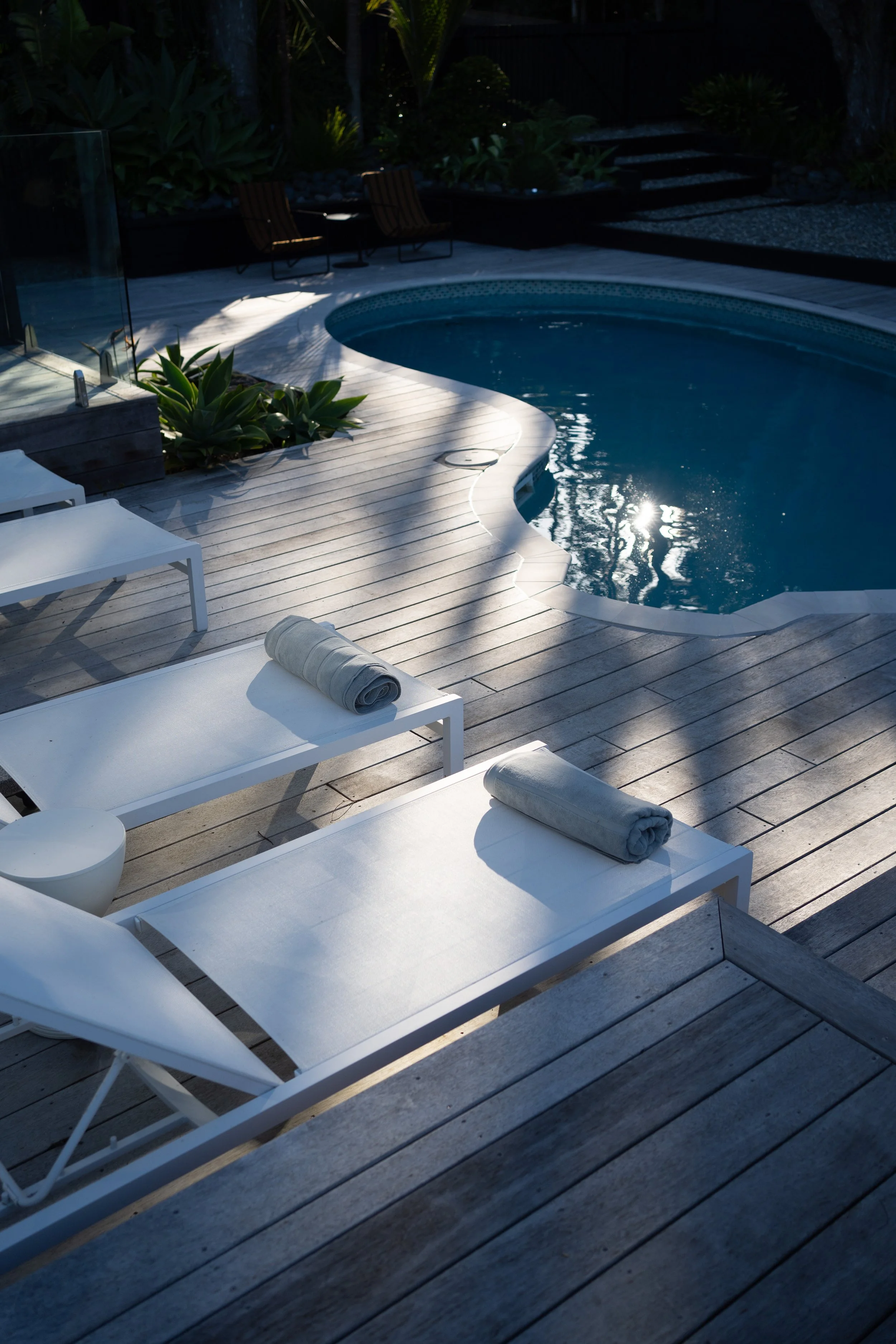 Empty outdoor pool area with white lounge chairs, towels, and lush green plants, partly shaded and reflecting sunlight.
