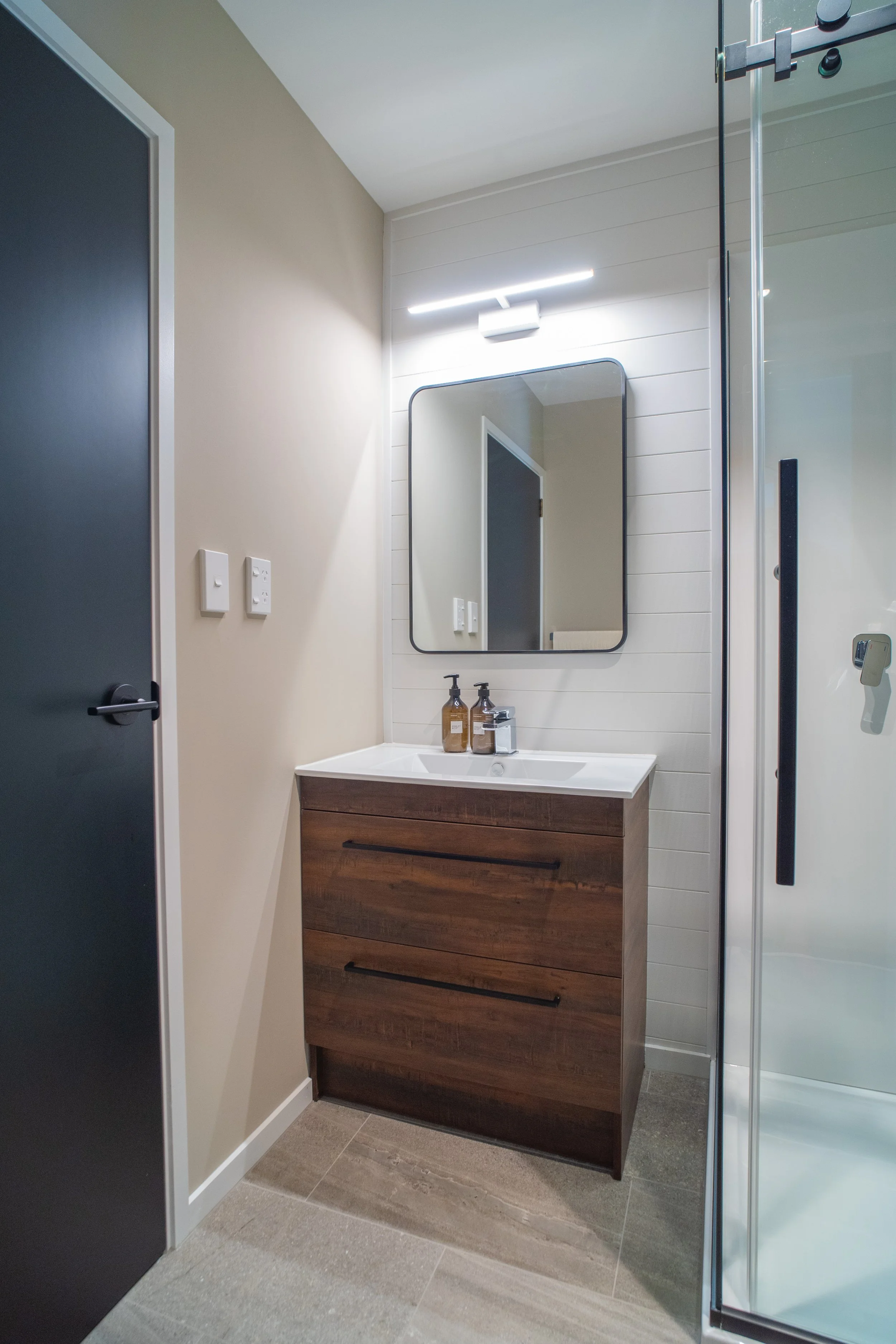 A modern bathroom with a small wooden vanity, a mirror, a shower with a glass door, and beige tiled flooring.