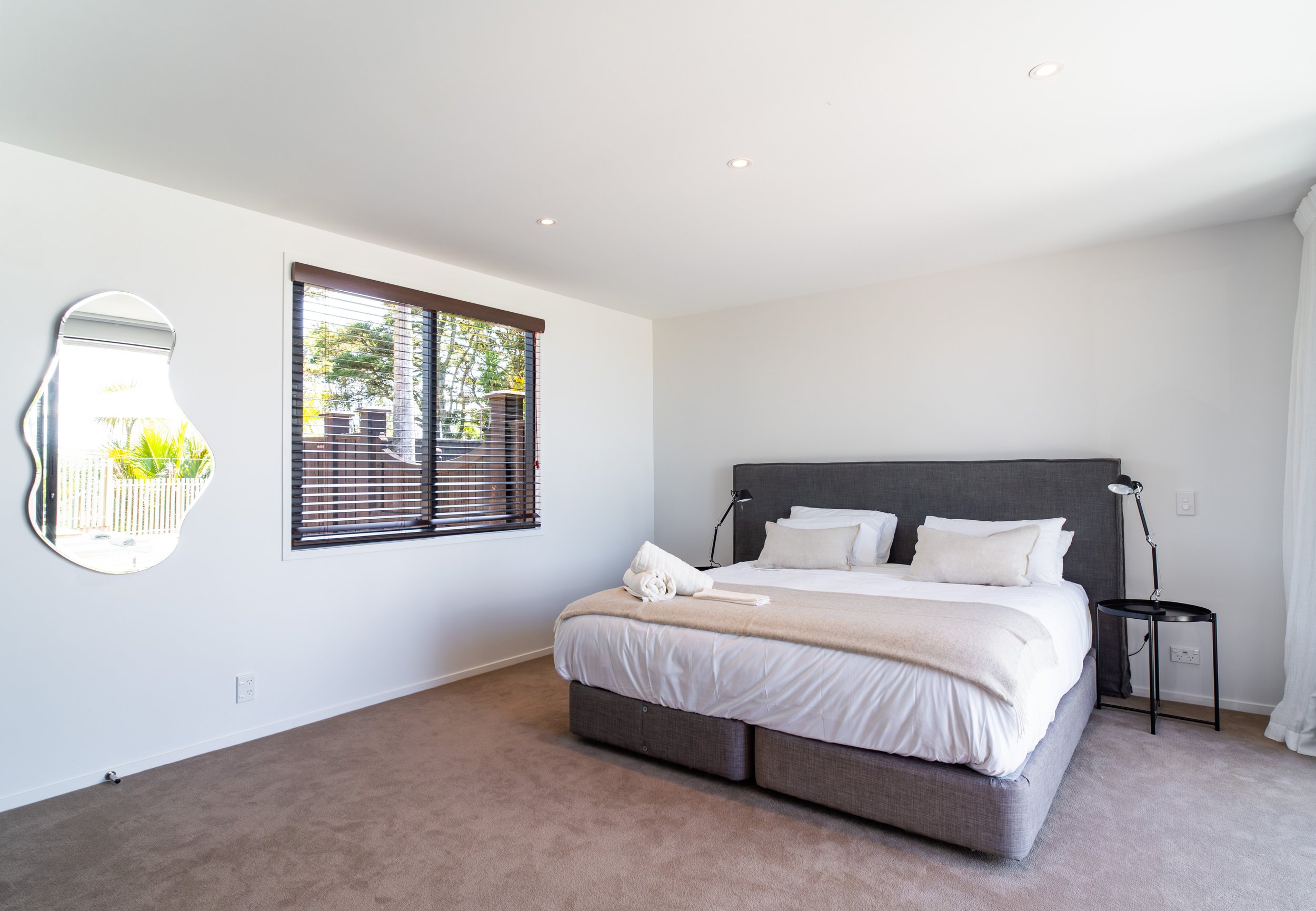 A bedroom with a large bed, grey upholstered headboard, white bedding, beige blanket, and pillows. Next to the bed are two black lamps on black bedside tables. There is a window with horizontal blinds and a unique shaped mirror on the white wall.