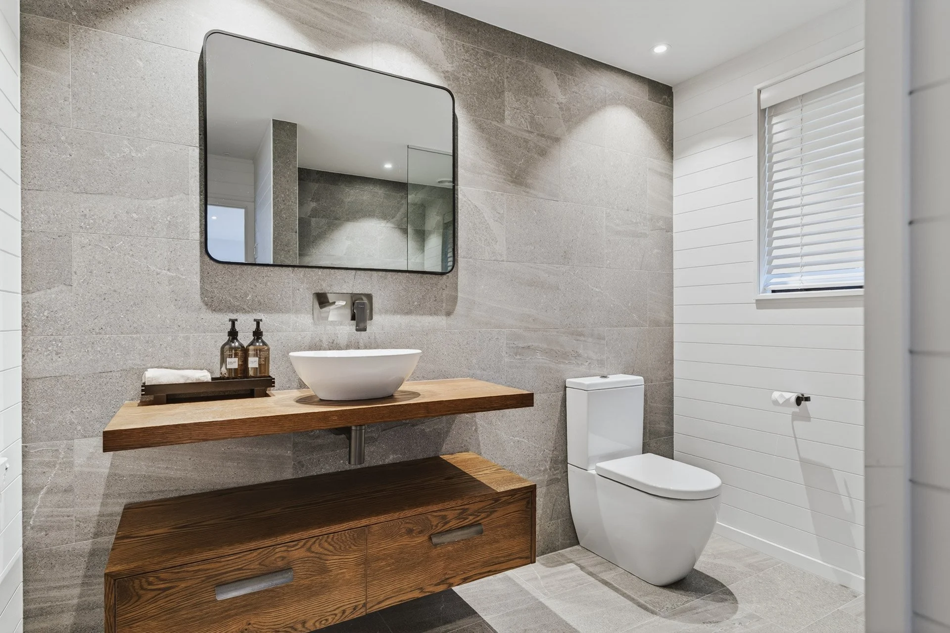 Modern bathroom with gray tiled walls, a white toilet, a wooden vanity with a vessel sink, a mirror, and a window with white blinds.