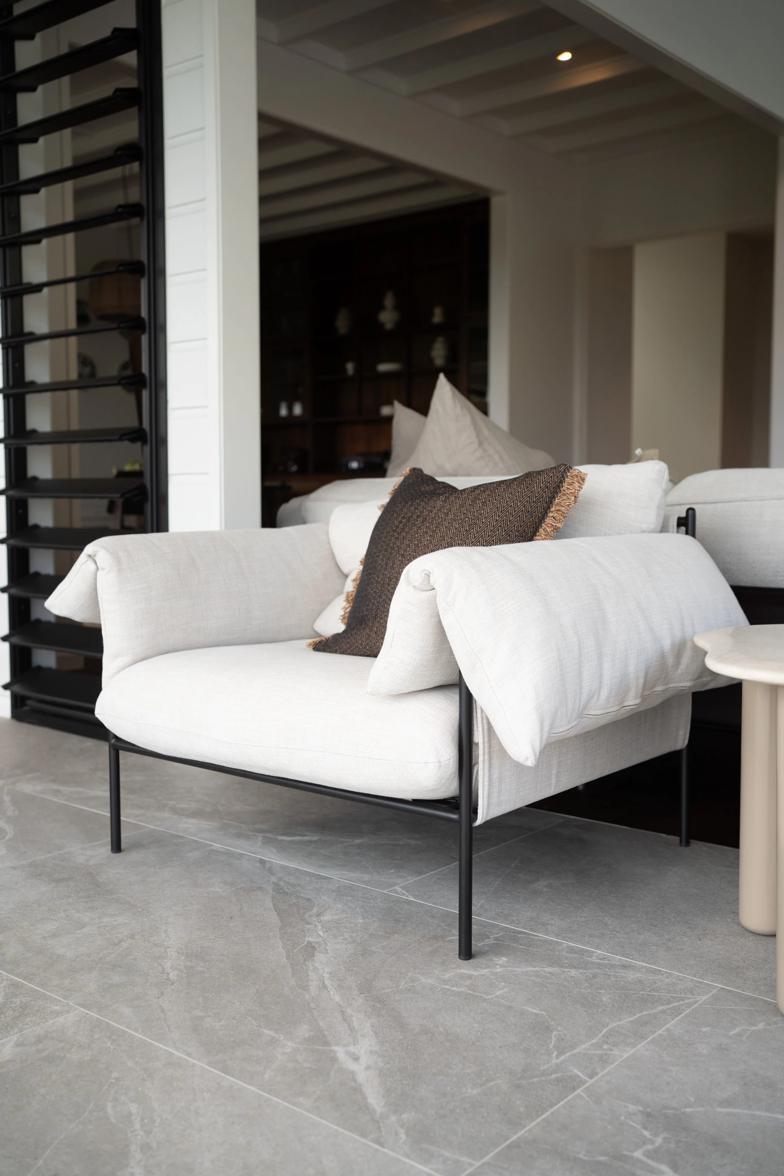 Living room with a white sofa and brown pillow, visible part of a small white side table, gray tiled floor, and a background with open doorway and dark wooden bookshelf.