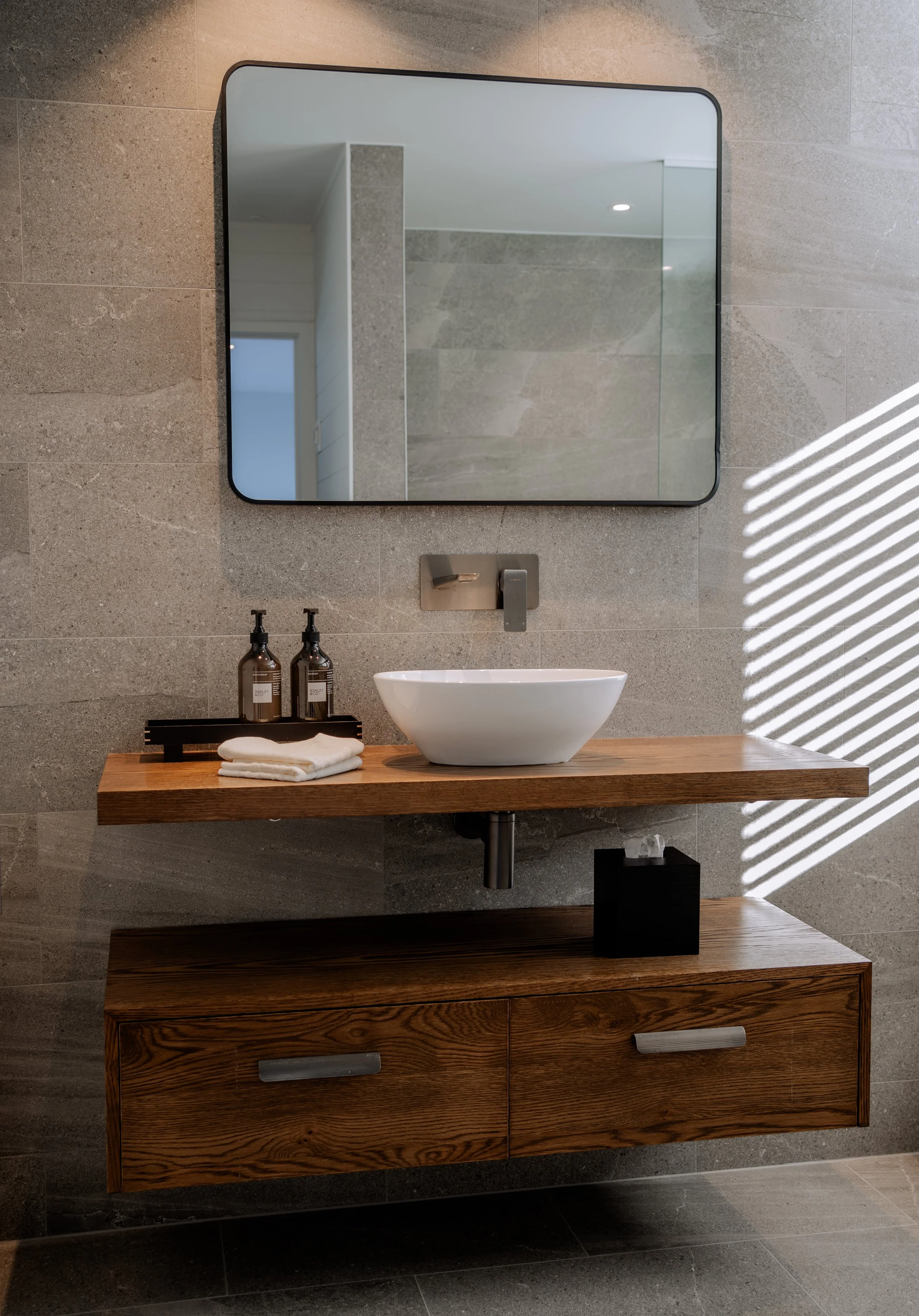 A modern bathroom vanity with a white vessel sink, a rectangular mirror, and brown bottles of soap or lotion on a black tray, all mounted on a wooden cabinet with drawers, against a gray tiled wall with sunlight streaming through a window.