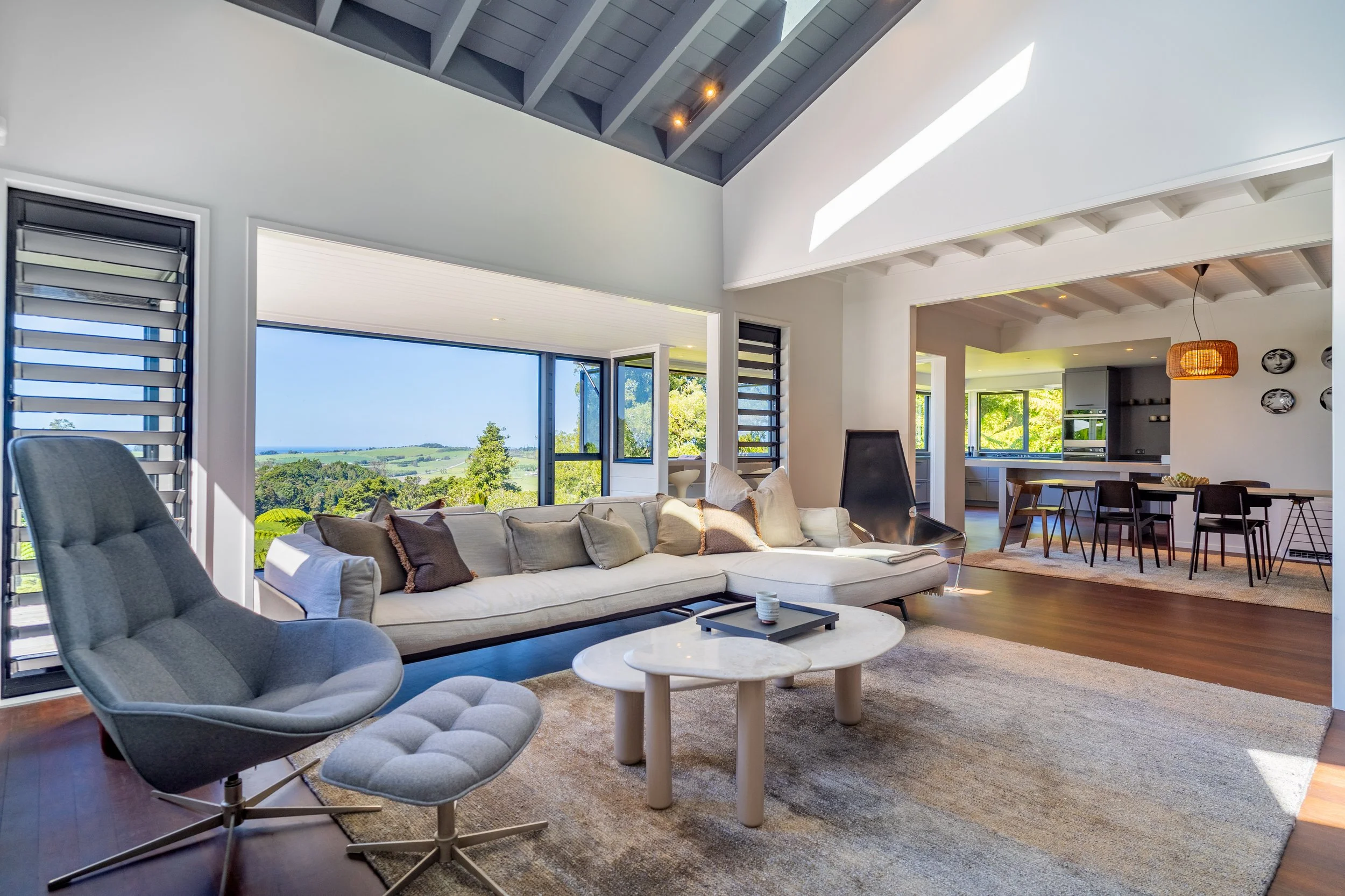 Modern living room with large windows showing a green landscape, gray armchair, and a white sectional sofa with beige and gray cushions, round marble coffee tables, and a textured beige rug.