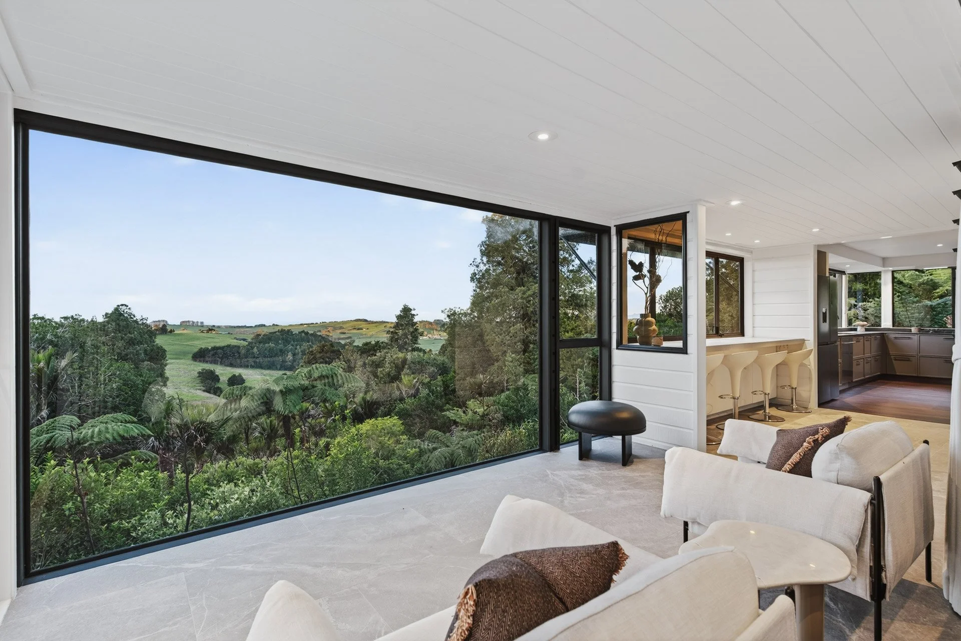 Living room with large glass window showing a scenic landscape of trees and rolling hills outside, modern kitchen with bar stools, white ceiling with recessed lighting, beige sofa with throw pillows.