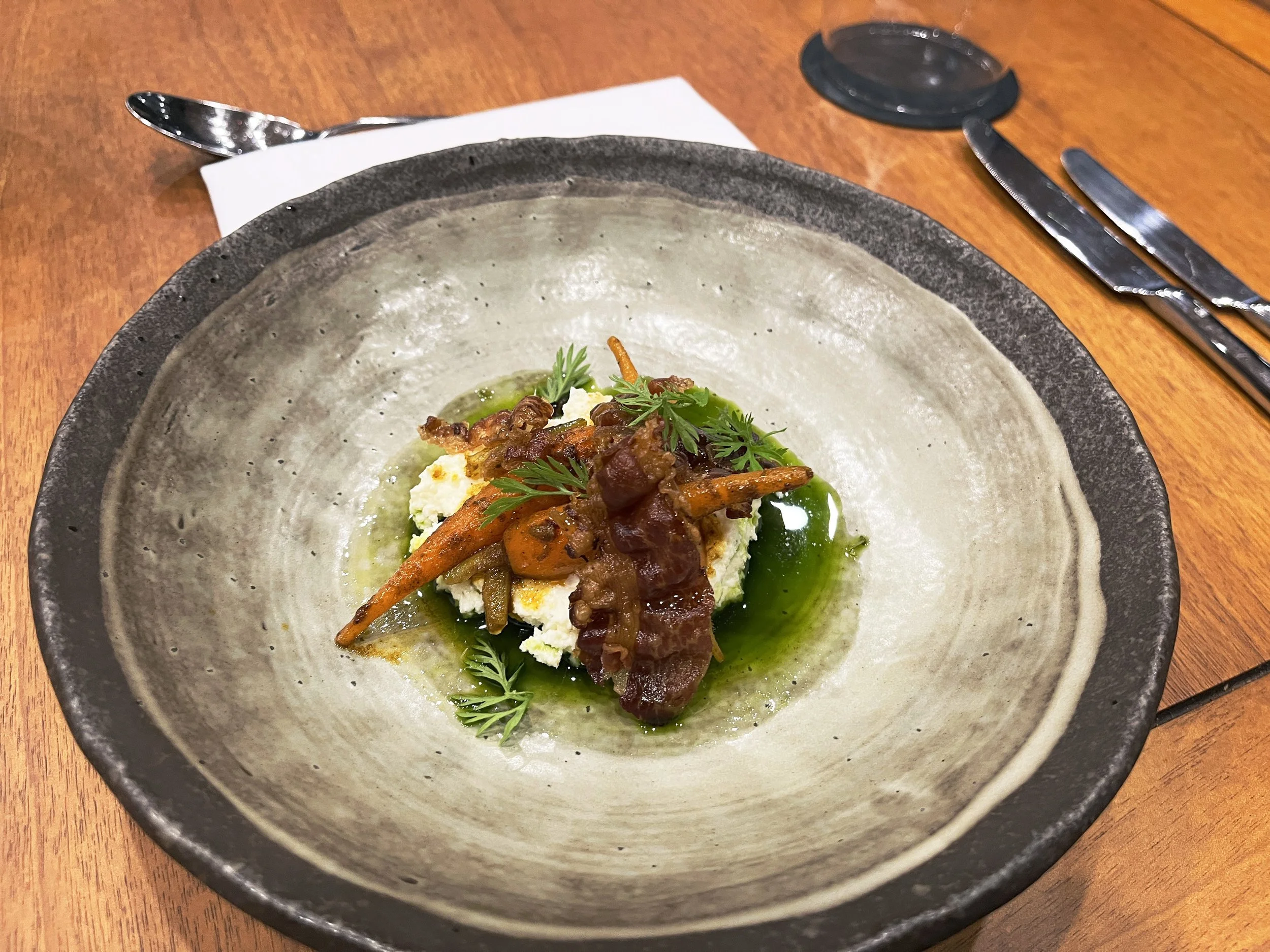 Plate of food with mashed potatoes topped with cooked vegetables and fresh herbs on a ceramic plate, placed on a wooden table with utensils and a napkin.