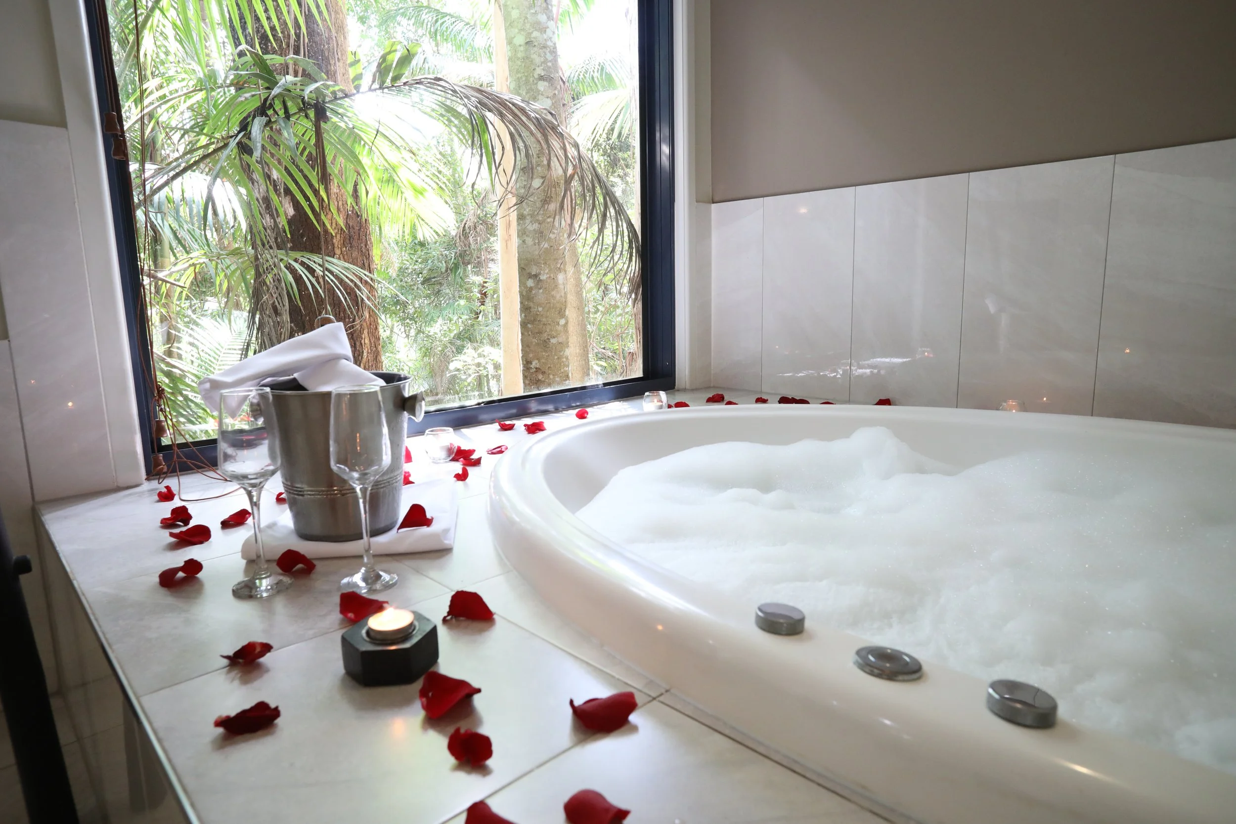 Spa bath set with rose petals, candles and champagne on ice, rainforest through the window