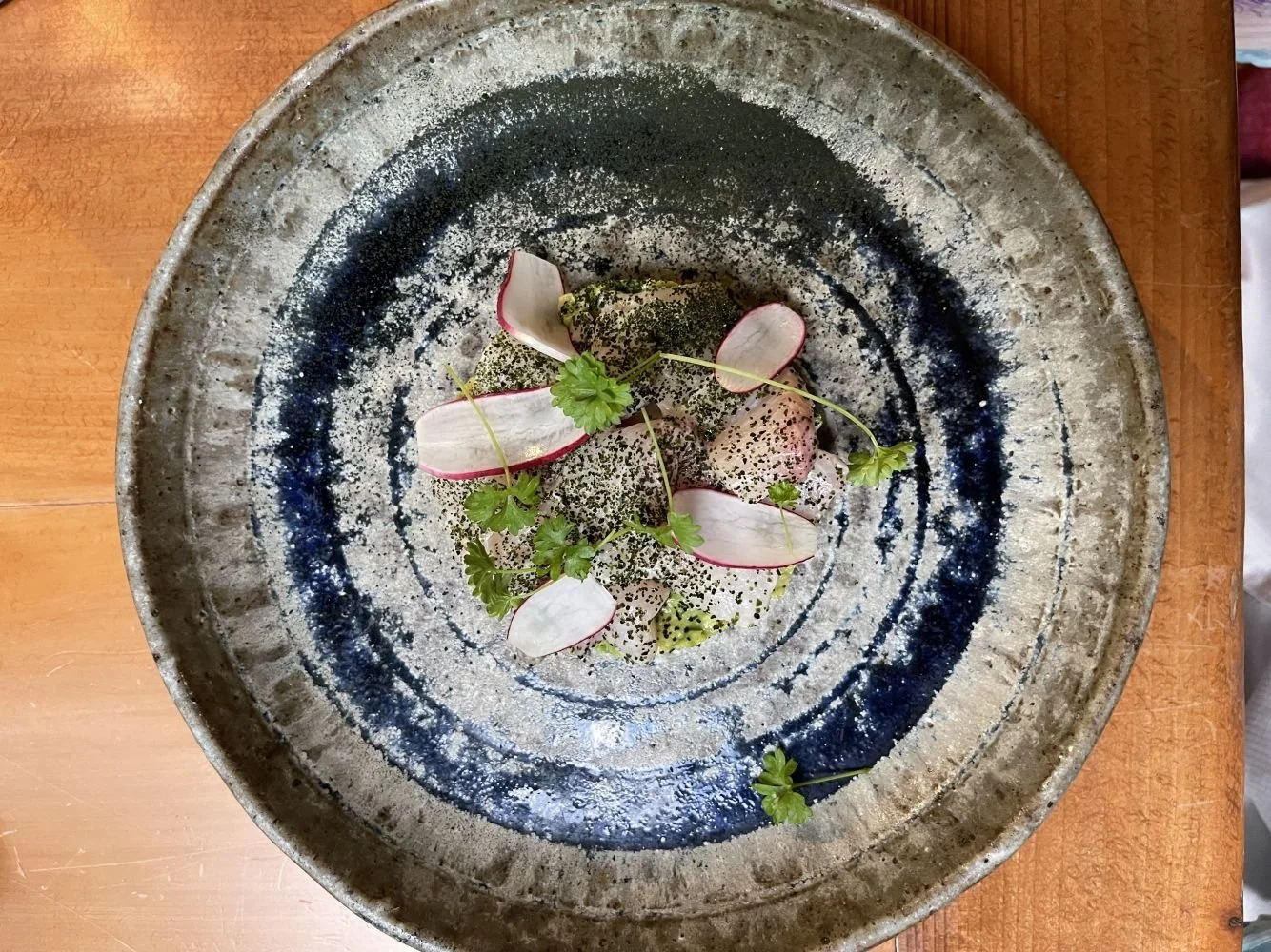 A palate of food with radish slices, microgreens, and black specks on a rustic ceramic plate on a wooden table.