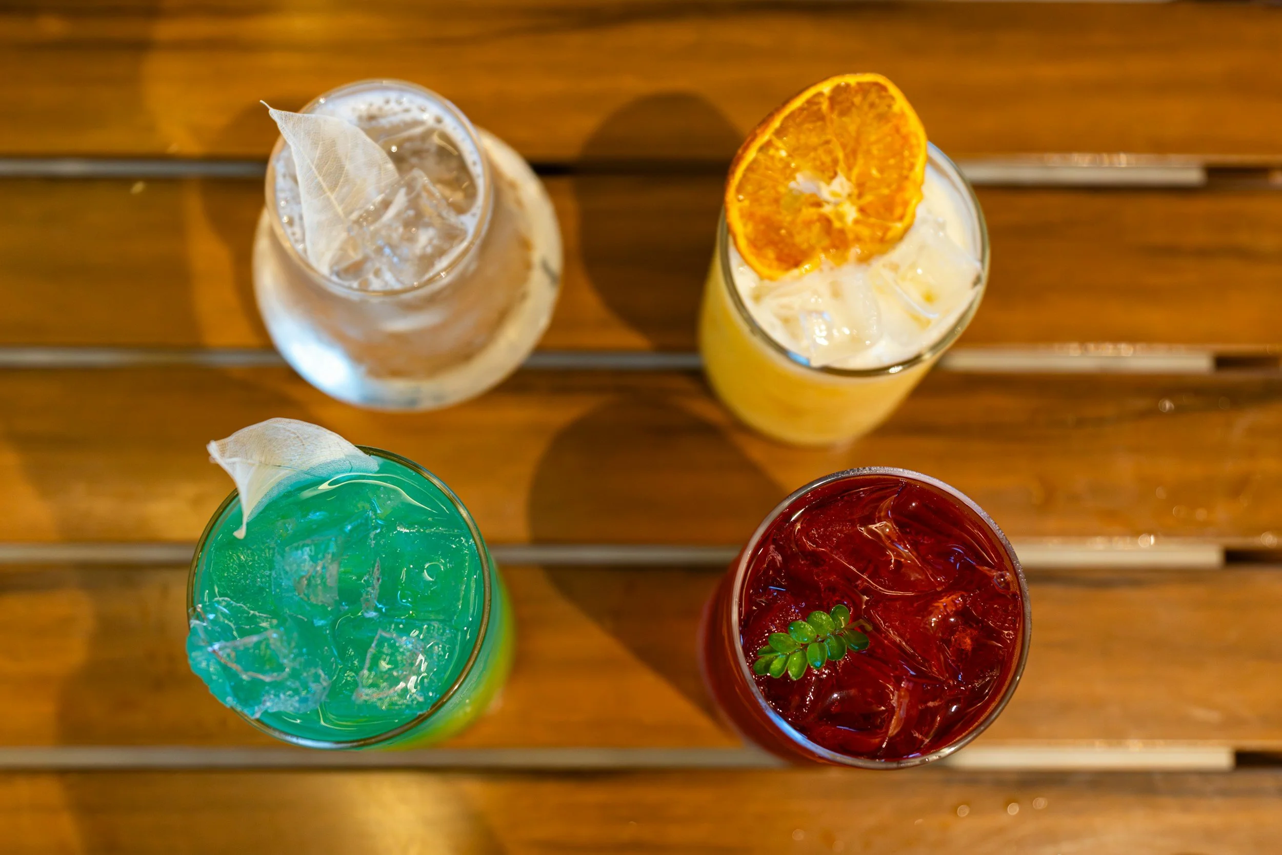 Top-down view of four colorful drinks with ice, garnishes, and slices of fruit on a wooden table.