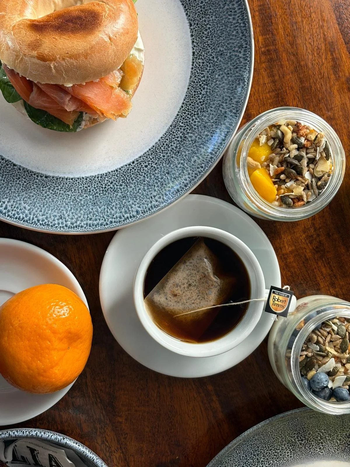A breakfast scene with a sandwich on a blue and white plate, a small jar of cereal and fruit, a cup of black coffee with a tea bag, an orange on a white plate, and a glass of water on a wooden table.