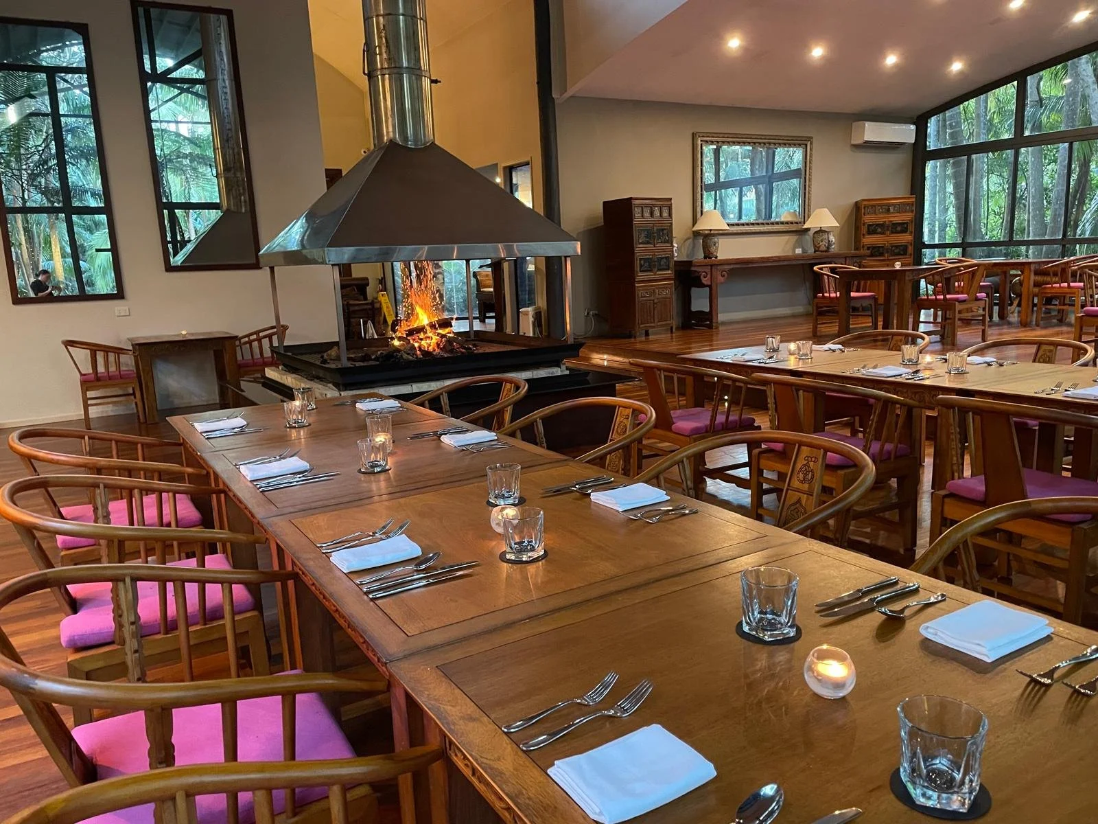Restaurant with wooden tables and chairs, a fireplace with a fire burning, large windows showing outdoor greenery, and table settings including plates, glasses, and silverware.