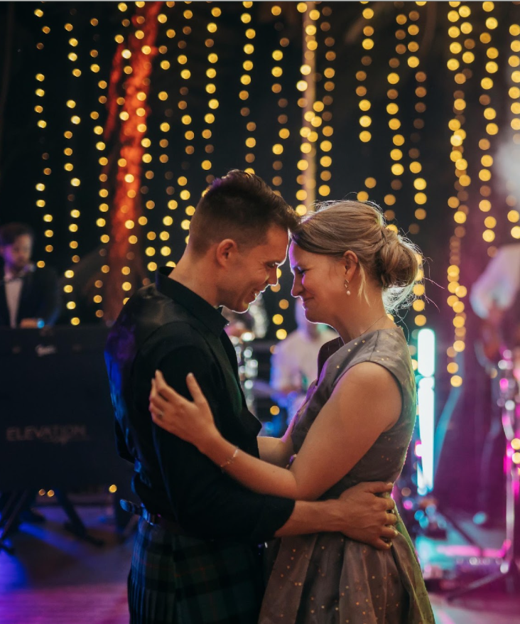 A couple dancing closely at a party or wedding reception, with string lights hanging in the background.