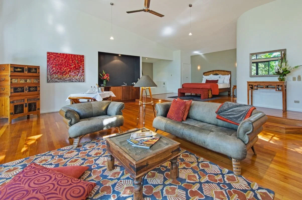A spacious living area with hardwood floors, a gray armchair, a gray sofa with red pillows, and a wooden coffee table. In the background, there's a bedroom with a bed and wooden furniture, and decorative artwork on the walls. The room is well-lit with natural light.