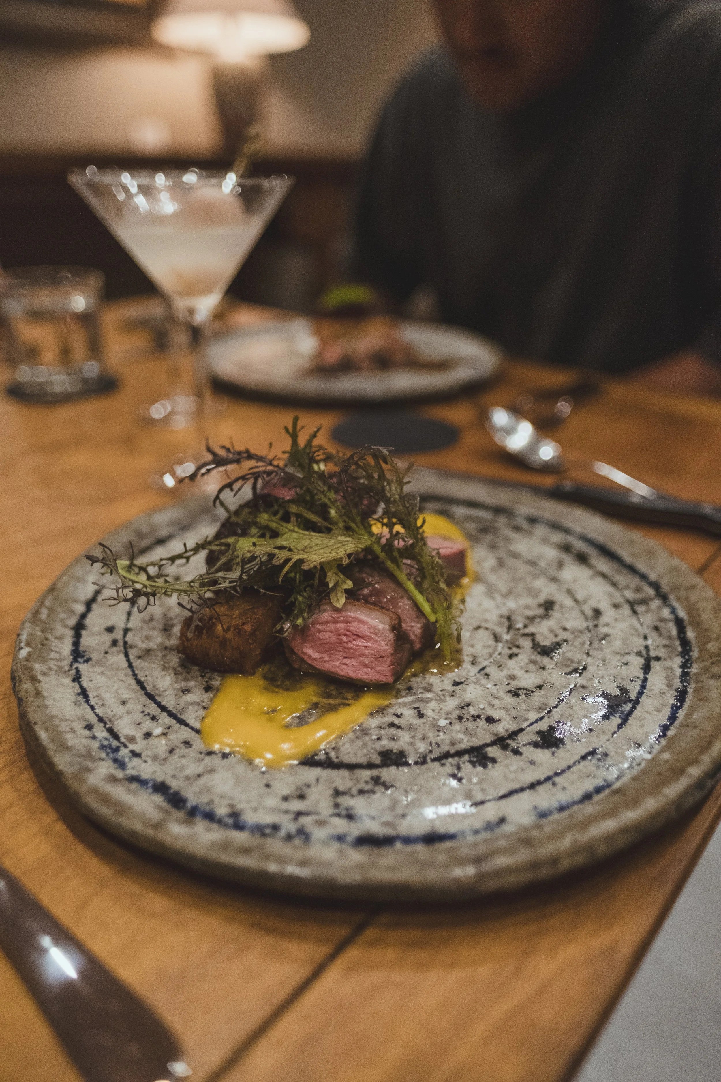 A close-up of a plated dish with slices of medium-rare steak garnished with herbs and a yellow sauce, on a speckled ceramic plate on a wooden table.