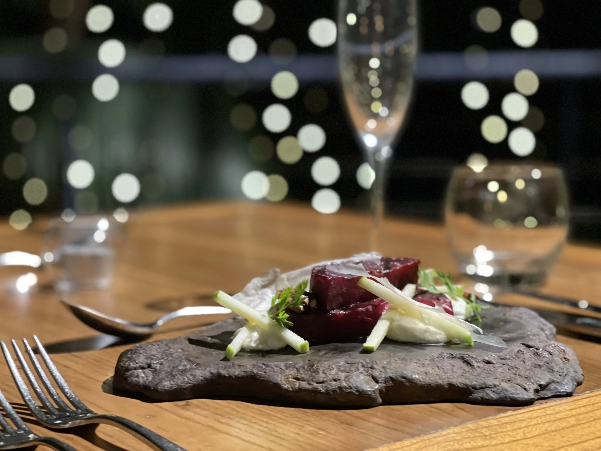 A close-up of a salad with beets, sliced cucumbers, and greens on a black plate on a wooden table, with a fork and knife nearby. In the background, there are blurred bokeh lights.