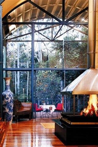 A cozy indoor space with large glass windows showing trees outside, a wooden floor, a fireplace with flames, a wooden table, red chairs, and decorative elements like a large vase.