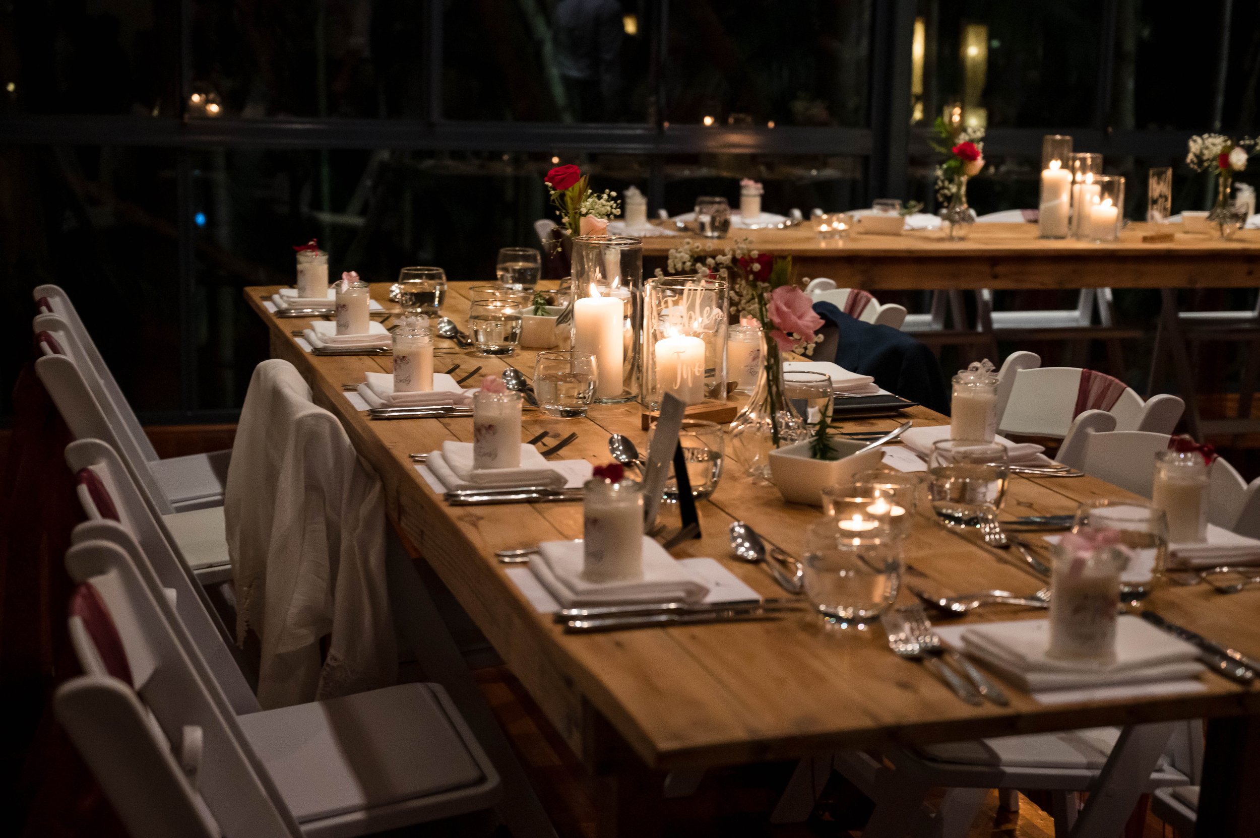 A long wooden banquet table decorated with candles, flowers, and tableware for a celebration or event in a dimly lit setting.