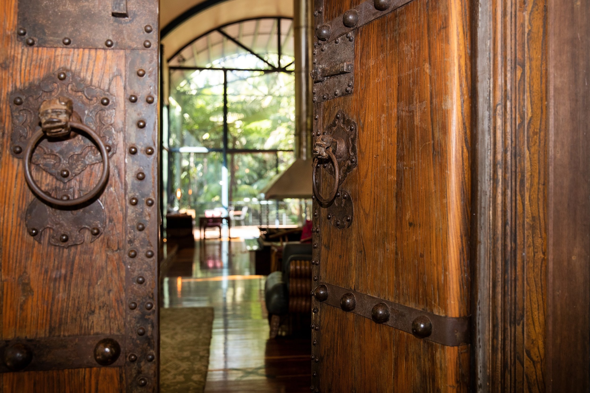 A view through a partially opened wooden door with metal hardware into a bright room with large windows and greenery outside.