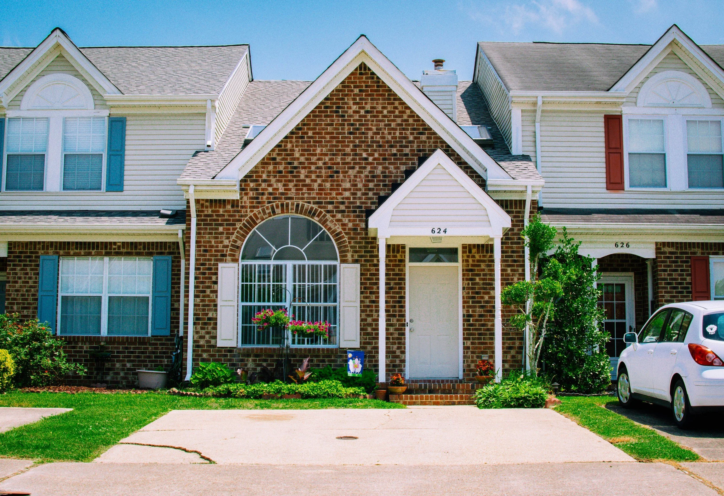 Front view of a two-story townhouse with brick and siding exterior, flower bed, and parked car nearby.