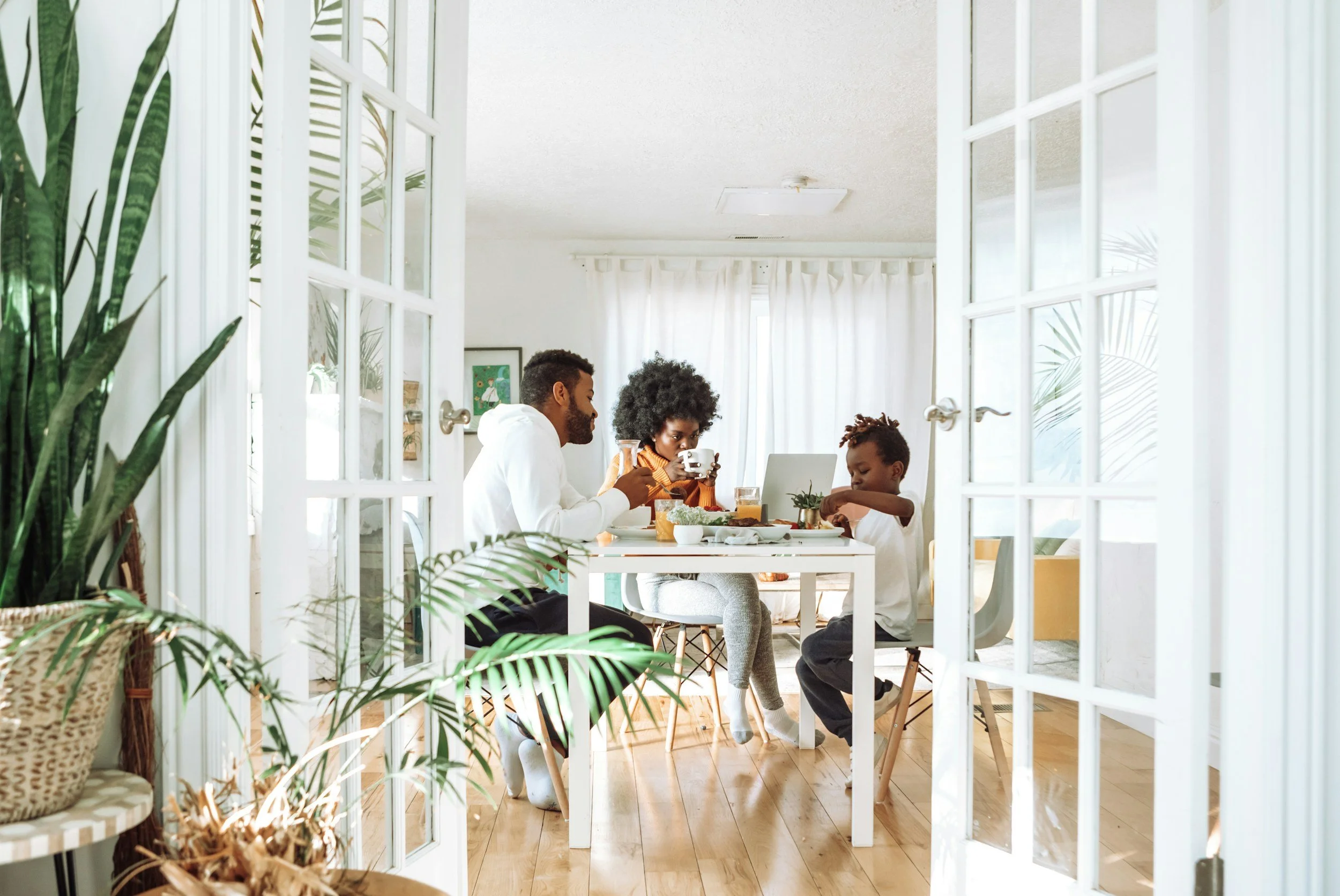 A family of three having breakfast or brunch inside a room, viewed through glass doors with white frames, with natural light coming in from windows and greenery outside.