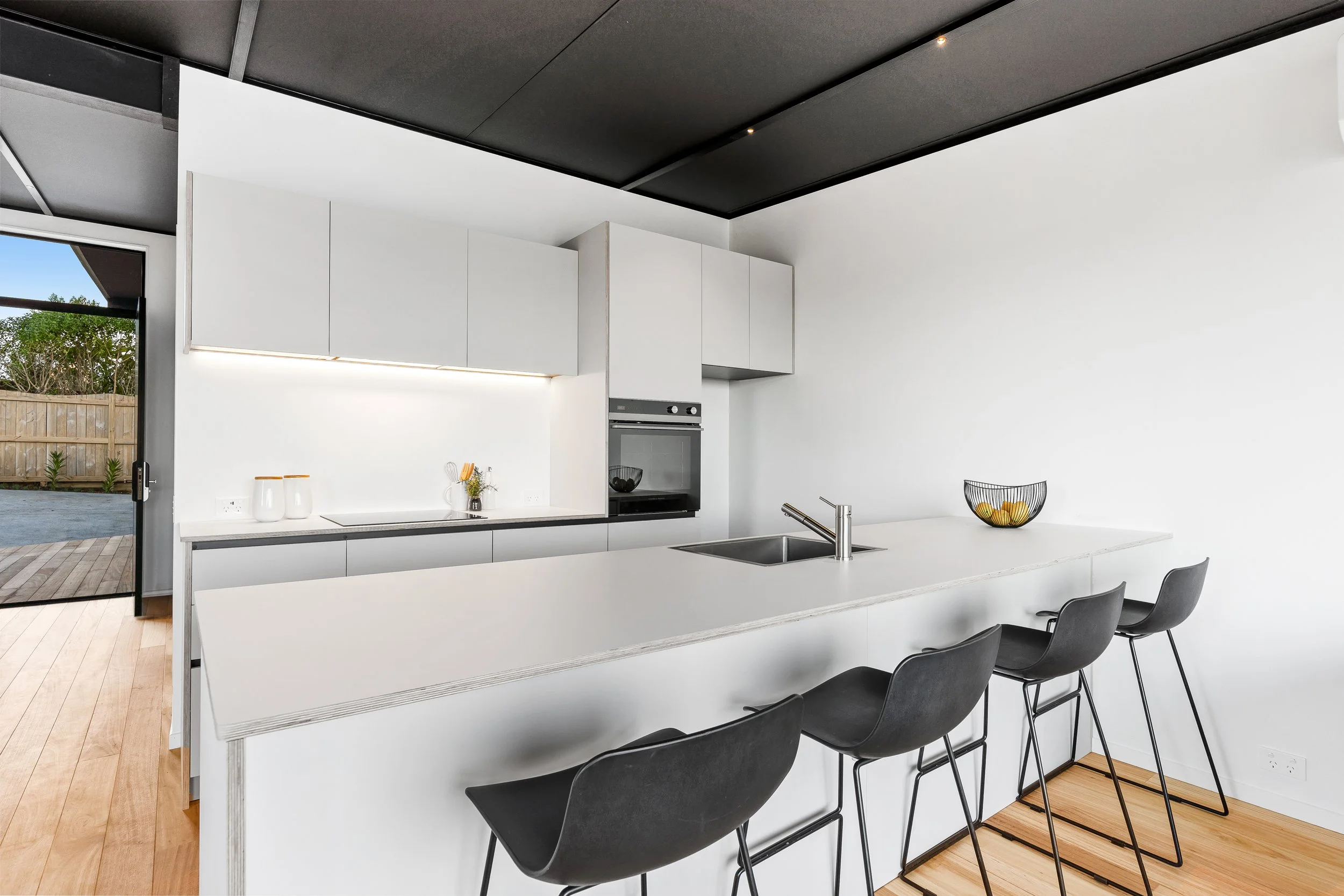 Modern minimalist kitchen with white countertops, black chairs, and upper white cabinets. The space features a built-in oven, a stainless steel sink, and represents new build projects plumbed by gravity plumbing