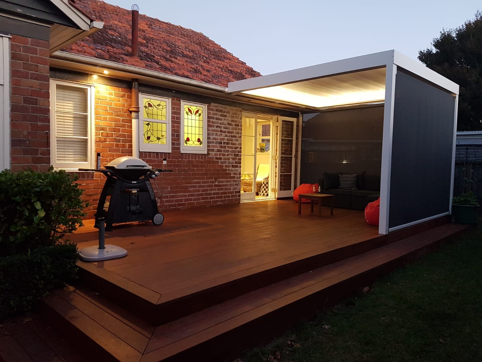 A cozy outdoor patio with a wooden deck, a barbecue grill, and seating area under a covered pergola attached to a brick house with stained glass windows, a renovation plumbing job by Gravity Plumbing
